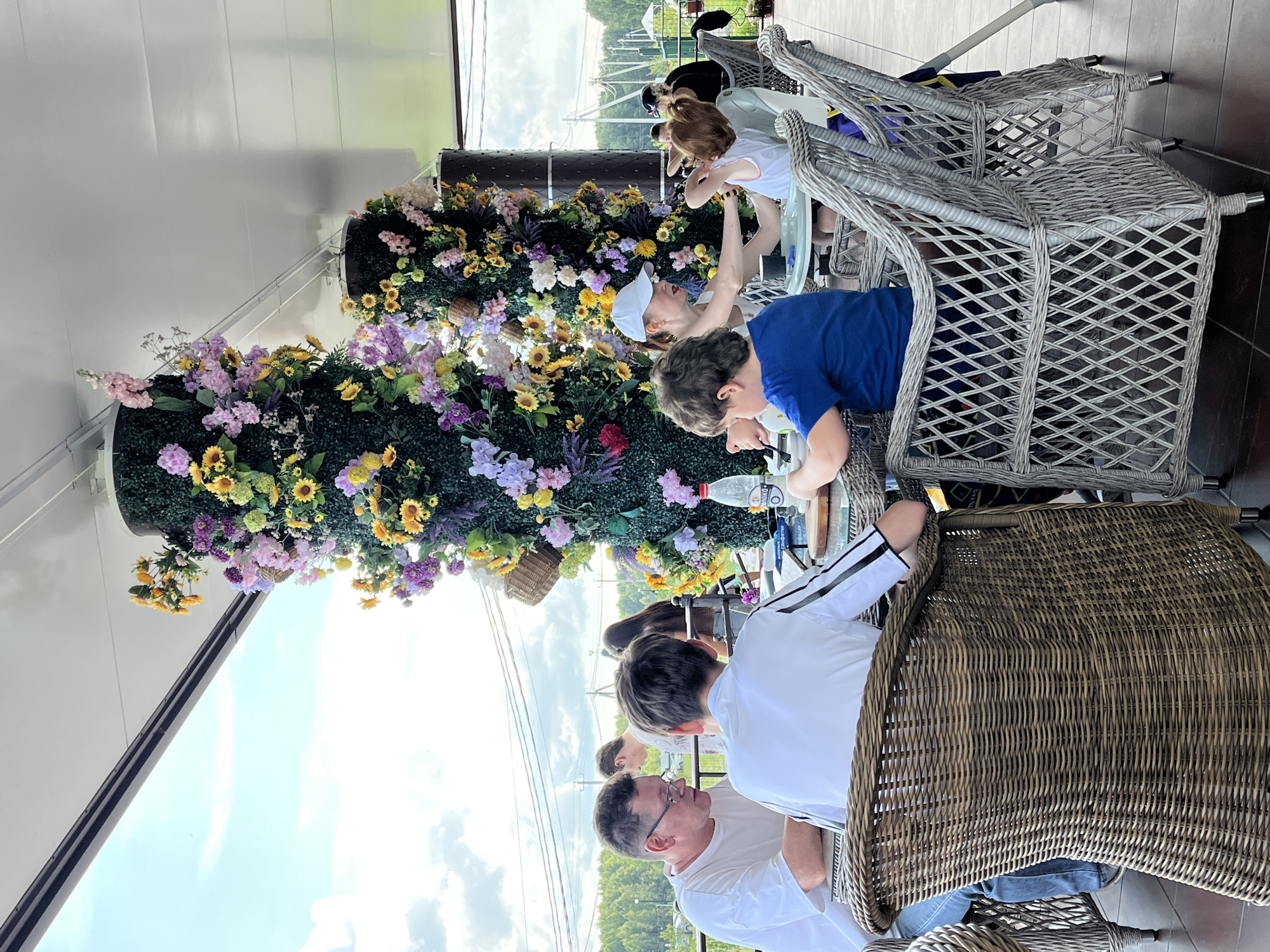 A group of people are seated at tables on an outdoor patio adorned with large, ornate floral columns. The patio is covered by a white roof structure, and in the background, a cloudy sky, trees, and distant structures are visible.
In the foreground, a man wearing a white t-shirt and glasses is seated at a table, engaged in conversation with a young person in a white shirt with black stripes. To their right, a boy in a blue t-shirt is seated at another table, with a woman in a white cap and tank top attending to a child with red hair.
The tables are covered with white tablecloths and set with plates and a bottle of water. The chairs are made of woven wicker. The floral columns are substantial, covered in dense greenery and decorated with a vibrant assortment of artificial flowers in various colors including purple, yellow, pink, and white. Some small wicker baskets are also attached to the columns.
The scene appears to be a leisurely gathering or meal at a restaurant or cafe. The overall atmosphere is relaxed and summery. The location is identified as Akhlebaikha (Kovriginskii S/S), Russia.