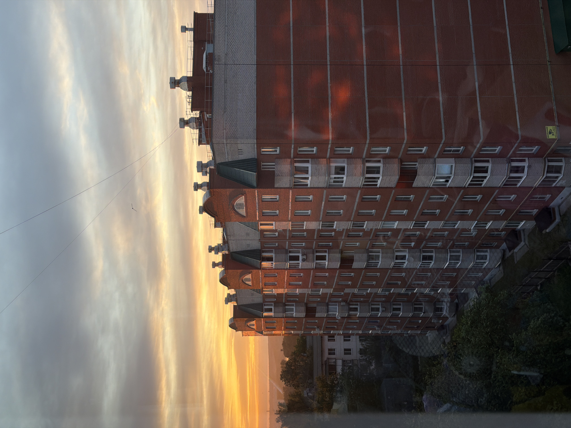 A tall, brick apartment building dominates the scene, with its facade reflecting the colors of the sunset. The building has multiple floors with rows of windows, some featuring balconies. A distinctive architectural design is visible on the top portion of the building. The sky is filled with shades of yellow, orange, and gray as the sun sets, casting a warm glow on the building. Below the apartment building, trees and other buildings can be seen, suggesting an urban landscape. The presence of a utility line crossing the sky is also noted. The building is likely in the city of Chaikovsky, Russia. There are no people visible in this image.
