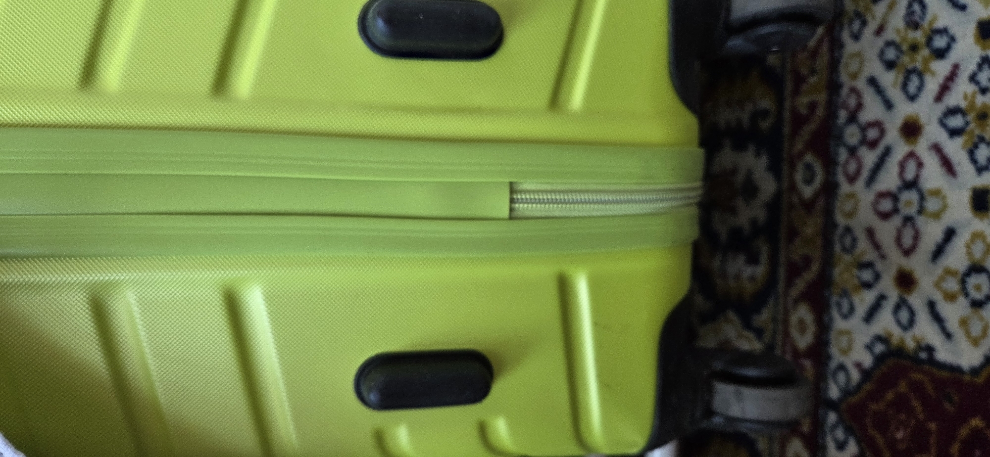 The image shows a close-up of a bright lime green suitcase on a patterned rug. The suitcase has a textured surface, a central zipper, and two black oval handles on the sides. The lower part of the suitcase reveals two wheels. The rug is dark with a repeating pattern of red, white, blue, and yellow geometric shapes and floral motifs. The scene is indoors, with no visible windows or external environment. The image does not contain any information that allows for the determination of its location in the city of Lipetsk, Russia, or any specific actions, interactions, or people.