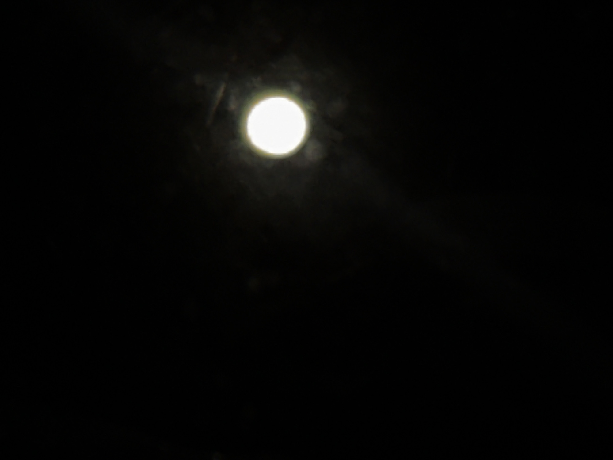 A full moon is visible in a dark night sky. The moon is a bright, circular object with a faint halo around it. The sky is mostly black, with some faint smudges and light scattering that may be dust or atmospheric conditions. There are no discernible features of the city of Zatoka, Ukraine, or any other location present in this image. No people, objects, actions, or interactions are visible.