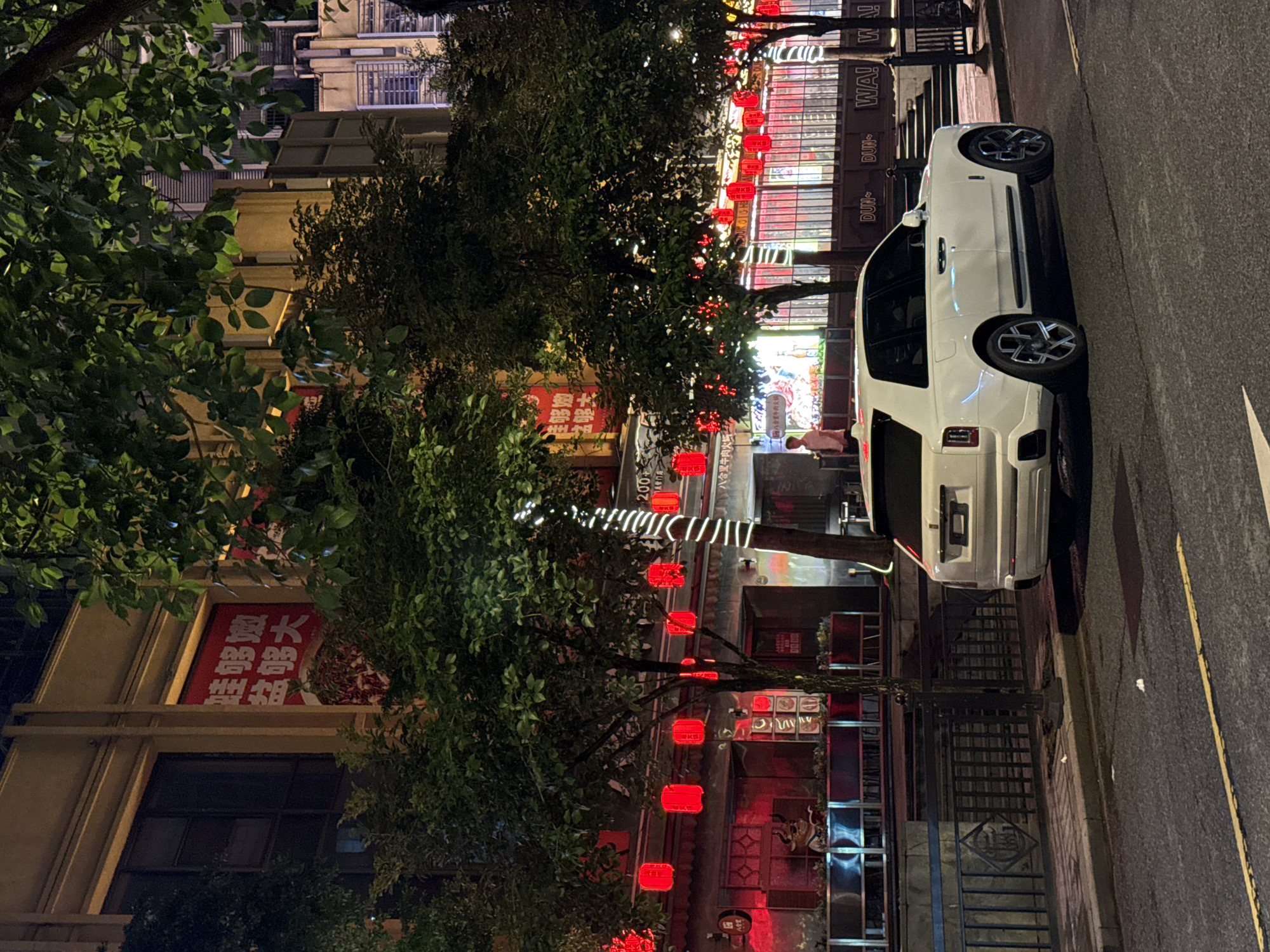 This media file depicts a street scene at night in Haizhu District, China. A white luxury SUV is parked on the right side of a street lined with trees. Red lanterns are hung from the trees, illuminating the foliage with a warm glow. The buildings on the left side of the street are adorned with neon signs and more red lanterns, suggesting a lively commercial area, possibly with restaurants or entertainment venues.
One of the buildings features a large red sign with Chinese characters, indicating a business or establishment. The street appears to be paved, with a yellow line marking the edge of the road. The time of day is clearly night, as evidenced by the artificial lighting and the darkness of the sky. The weather appears to be calm, with no indication of rain or strong winds.
A person is visible on the sidewalk near the building, dressed in a pink shirt. The overall atmosphere is vibrant and bustling, characteristic of a lively urban night scene in China. The presence of the luxury car and the decorative lighting hints at a prosperous and dynamic environment.