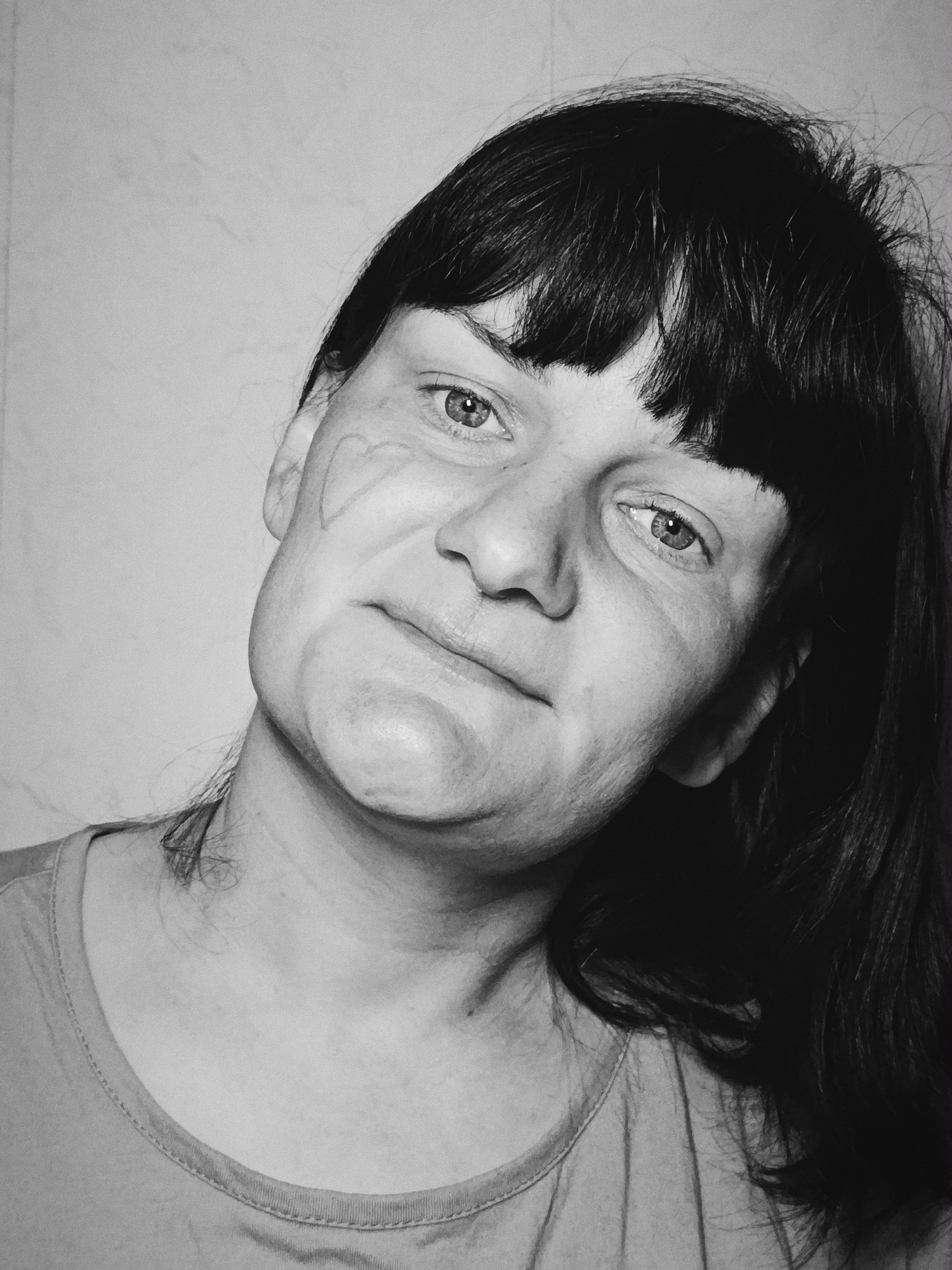 A black and white headshot features a middle-aged woman with dark hair and bangs. Her face is slightly turned, and a faint impression or mark is visible on her left cheek. The background is a plain, light-colored wall. She is wearing a simple t-shirt. No location of Фоки, Russia, is evident in the image.