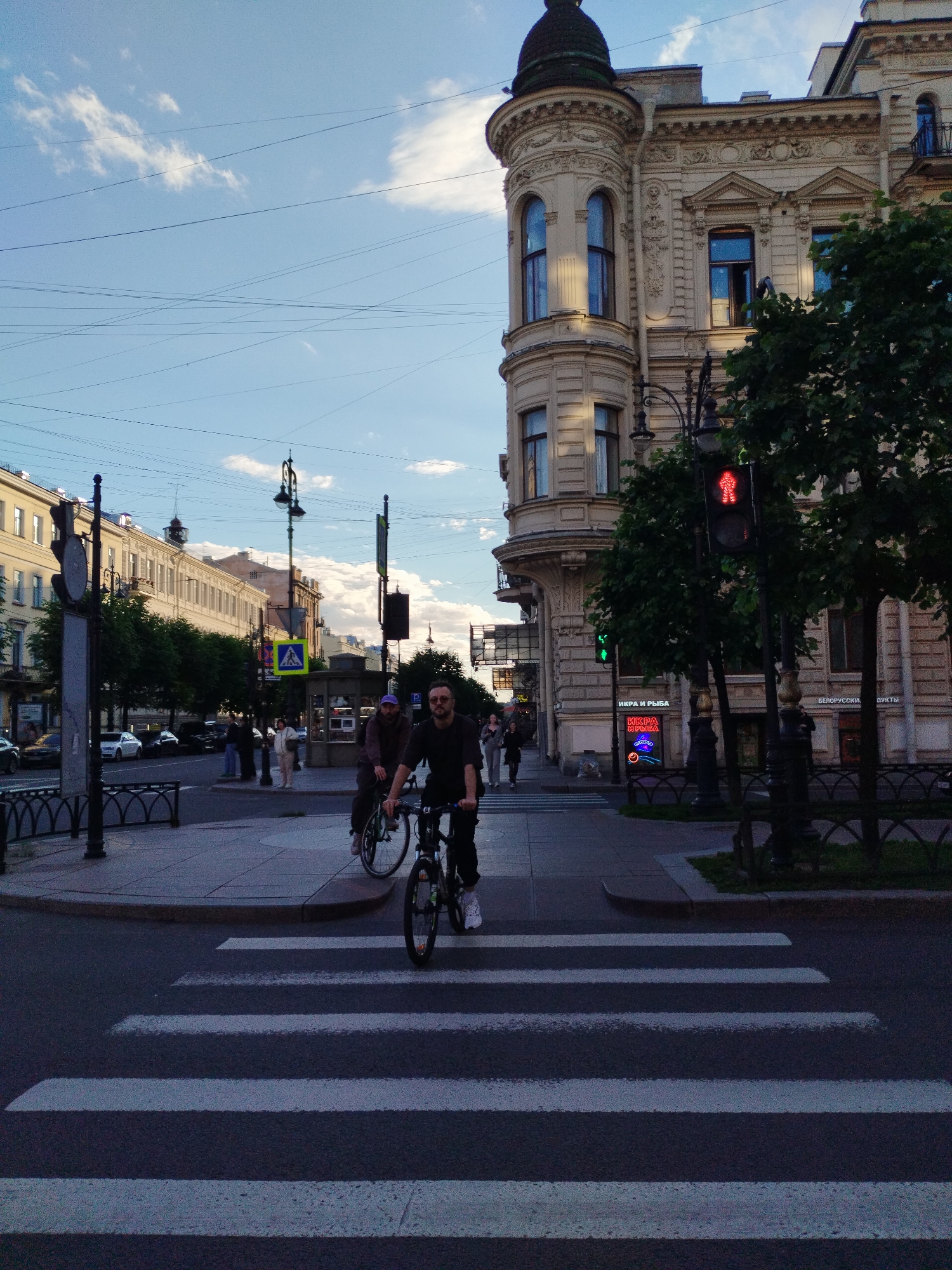 A street scene in Saint Petersburg, Russia, features a pedestrian crossing in the foreground with two men on bicycles approaching it. The man in the foreground wears sunglasses and a dark shirt, while the man behind him wears a purple cap and a brown hooded jacket. They are on bicycles with visible wheels and frames. To the right, a traffic light displays a red pedestrian signal. Behind the crossing, a sidewalk is lined with trees and ornate buildings. One building, on the corner, has a distinctive turret and decorative facade. Further down the street, more buildings and trees are visible. The sky above is blue with scattered white clouds, and numerous electrical wires crisscross the upper part of the image. The overall lighting suggests it is late afternoon or early evening.