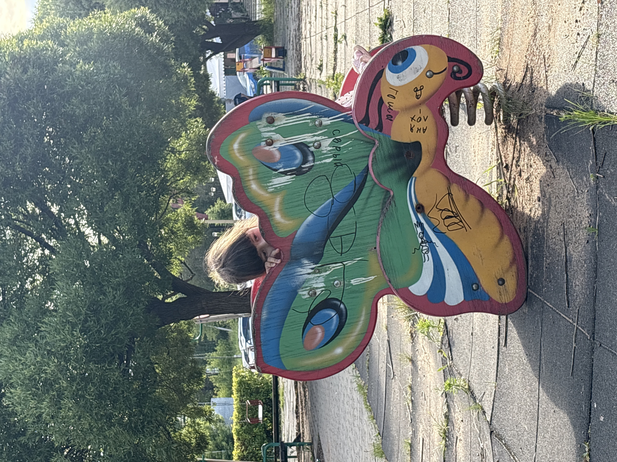 A young person, with long brown hair, peers over a colorful butterfly-shaped playground ride. The ride is made of wood and painted with bright green, yellow, red, and blue colors, depicting a whimsical face with a large blue eye. There are visible scratch marks and some graffiti on the ride. The person's face is partially obscured, but they appear to be looking down at the ride.
The setting is an outdoor playground with a paved surface of interlocking rubber tiles and some sparse grass growing in the cracks. In the background, there are trees with lush green foliage and playground equipment, including swings and a red seat. The lighting suggests it is daytime, likely late afternoon given the shadows. The overall mood is casual and playful.
Visible text on the ride, written in Cyrillic script, includes "Гаша" and "серый", and possibly other illegible markings. The location context is Chaikovsky, Russia.