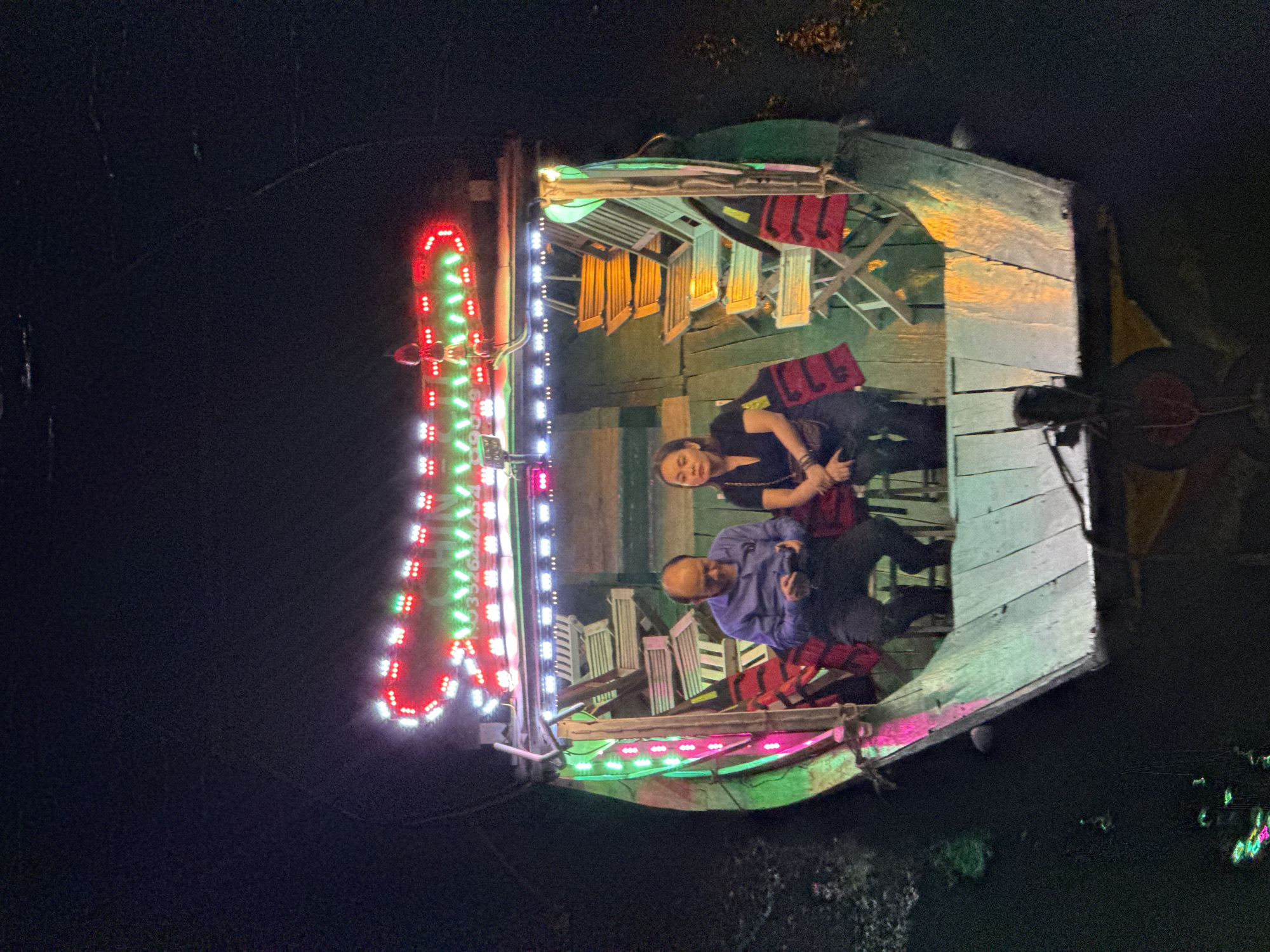 A nighttime, high-angle shot captures a long, narrow wooden boat illuminated by colorful LED lights, traveling on dark water, possibly in Hoi An, Vietnam. Two individuals, a man and a woman, are seated in the boat's middle section. The man, wearing a blue shirt and dark pants, is looking down at his phone. The woman, in a black top and dark pants, is looking forward. Life vests, red and with white lettering, are visible behind them and also on some of the empty, folded chairs stacked within the boat. The boat's exterior is painted green, with the wooden interior showing signs of wear and tear. The water's surface reflects the boat's lights, with smaller lights also visible in the water, possibly from other boats or aquatic life. The scene suggests a leisurely boat ride, potentially a tourist activity, in a dimly lit urban environment.