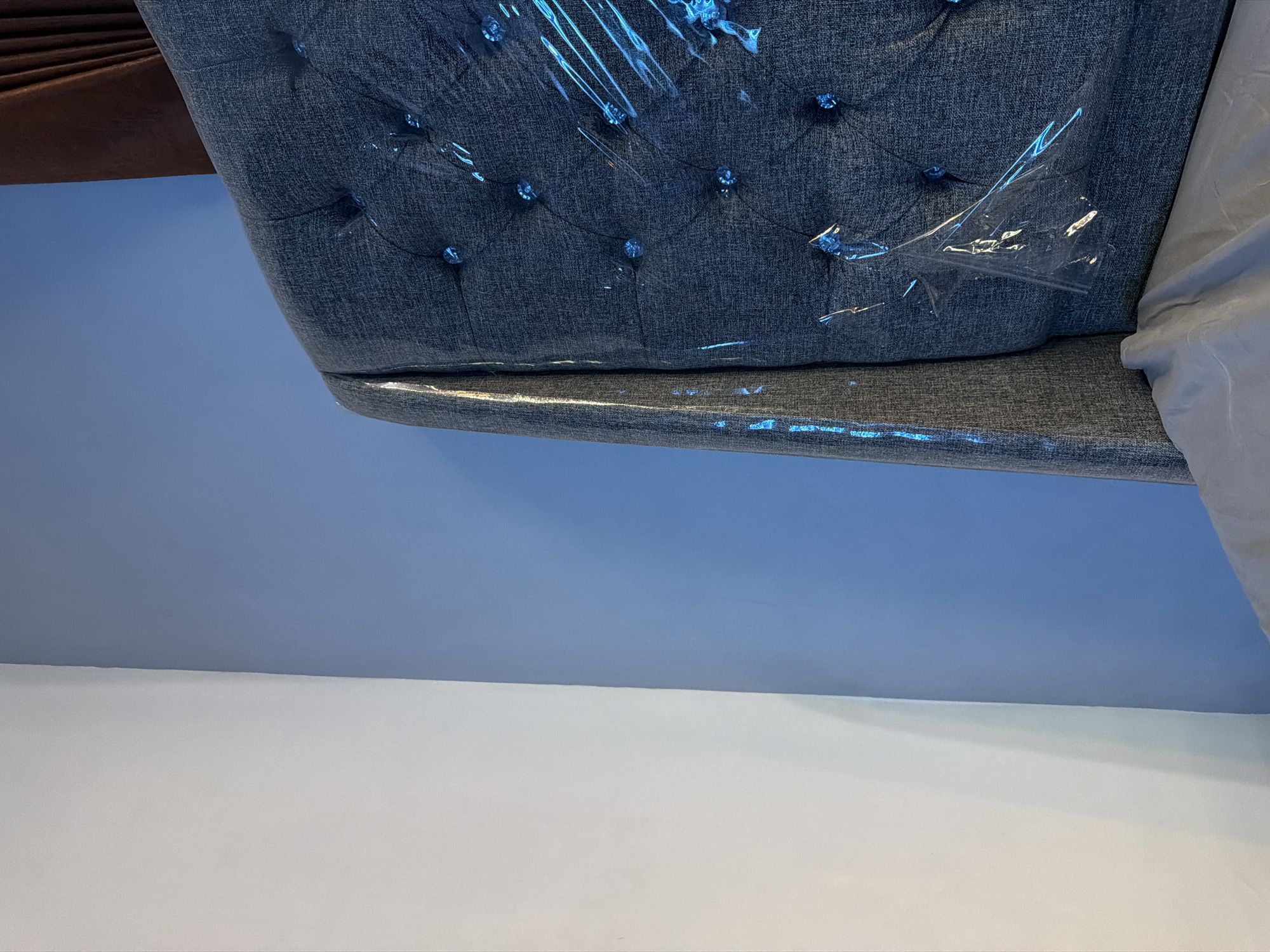A close-up, head-on view captures the corner of a tufted, dark blue headboard, still partially wrapped in clear plastic. The headboard features diamond-shaped tufting with small, faceted blue embellishments at each intersection. The plastic wrap catches the light, creating streaks of blue and white reflections. To the left of the headboard is a wall painted a soft, light blue, which transitions to a stark white wall on the far left of the frame. A portion of a dark brown curtain is visible in the upper right corner. The overall scene suggests a room that is either newly decorated or recently prepared for storage, with a focus on the textured headboard and the contrast of the adjacent walls. The image provides no discernible information about the city of Wonoayu, Indonesia.