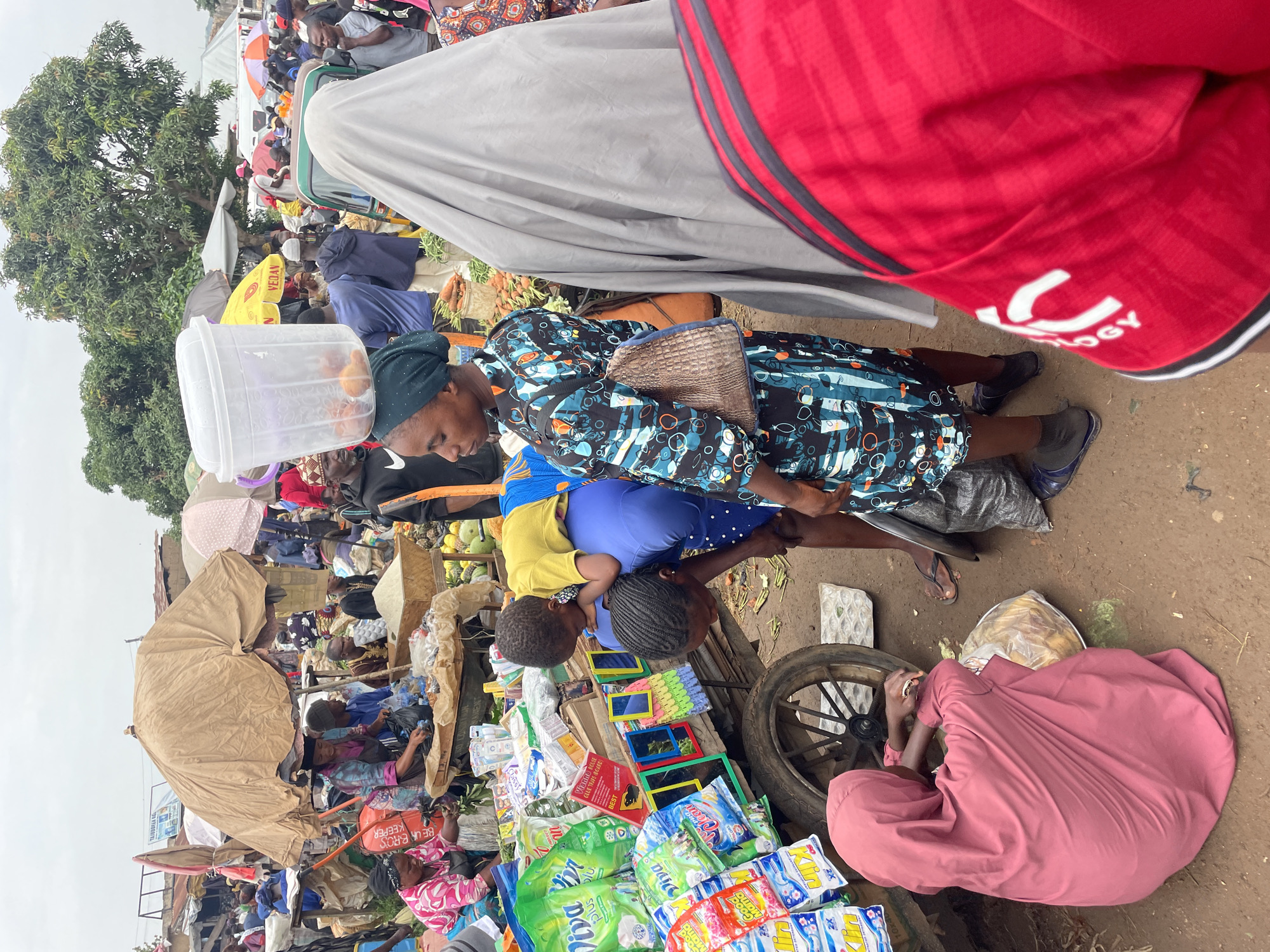 The image depicts a bustling outdoor market scene in Gura Top, Nigeria. In the foreground, a woman dressed in a patterned blue and black dress and a headscarf is carrying a child on her back. The child, wearing a yellow shirt, is leaning forward, resting their head on the woman's shoulder. A large clear plastic container filled with fruit is balanced on the woman's head. Next to them, another woman in a pink hijab is kneeling on the ground, tending to a plastic bag of produce. Behind them, a cart is laden with various packaged goods, including detergent. The market is crowded with people, many under makeshift awnings and umbrellas, suggesting it is an overcast day. Stalls display a variety of items, including produce and packaged goods. The ground is dusty and appears to be made of dirt. In the background, more stalls and people are visible, along with trees and some buildings. The overall impression is one of vibrant commerce and daily life in a Nigerian market.