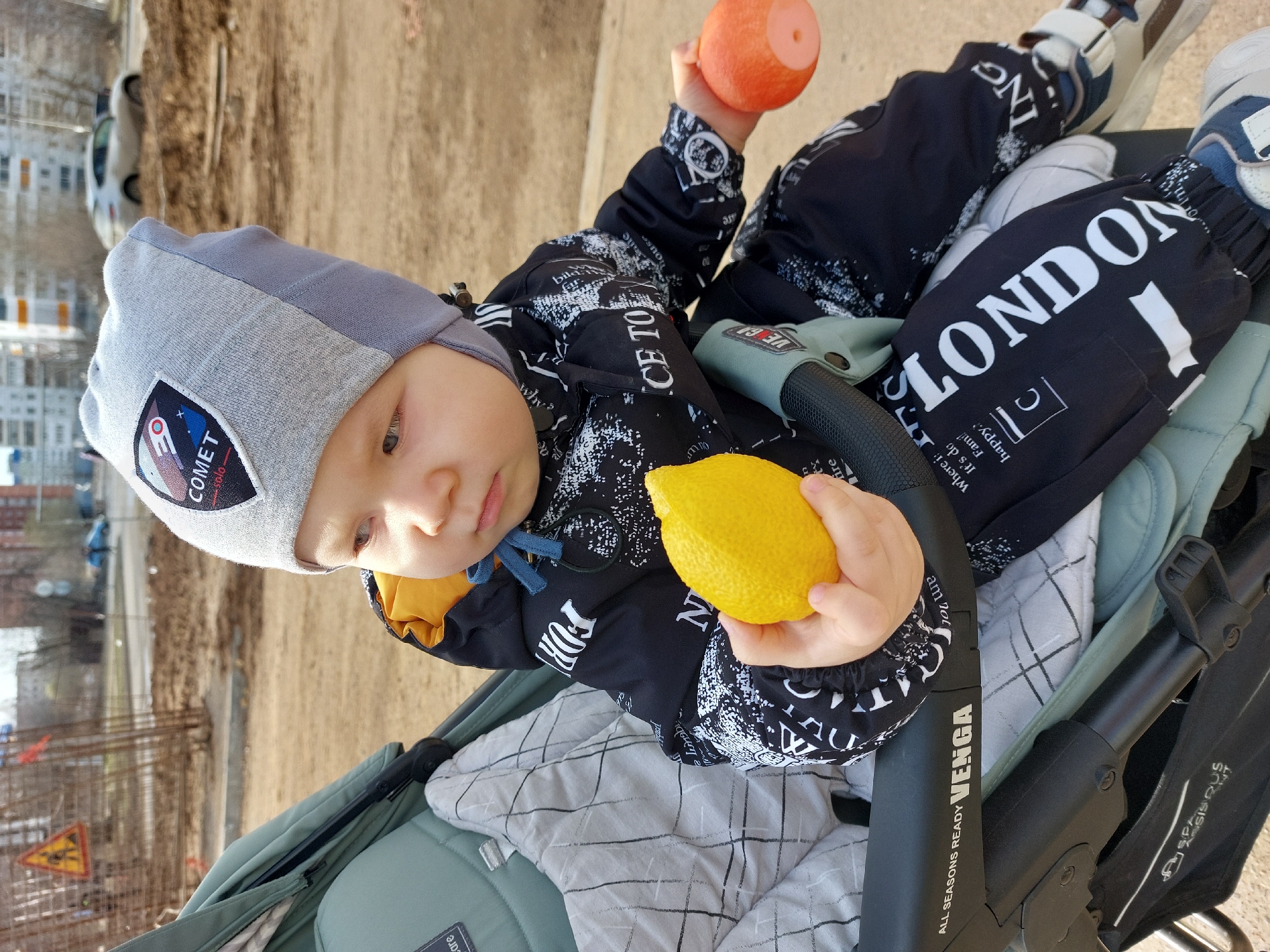 A child in a grey hat and black jacket with white lettering, is seated in a VEGA stroller, holding a yellow lemon in their right hand and an orange sphere in their left. The child's pants are black with "LONDON" printed in large white letters. The background shows a construction site and buildings. The image was taken in the city of Chaikovsky, Russia.