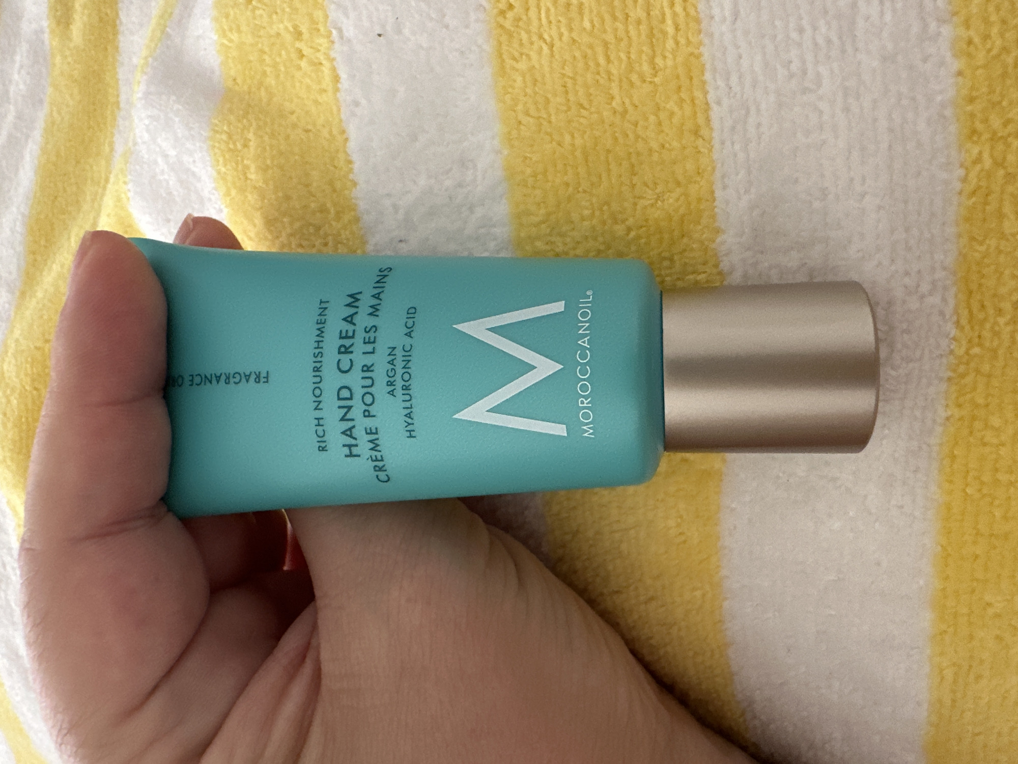 A hand holds a teal tube of Moroccanoil hand cream with a silver cap. The tube is upright and the hand is gripping the top portion. The background is a yellow and white striped terrycloth towel. The scene is indoors. The image does not provide any information about the city of Krefeld, Germany.