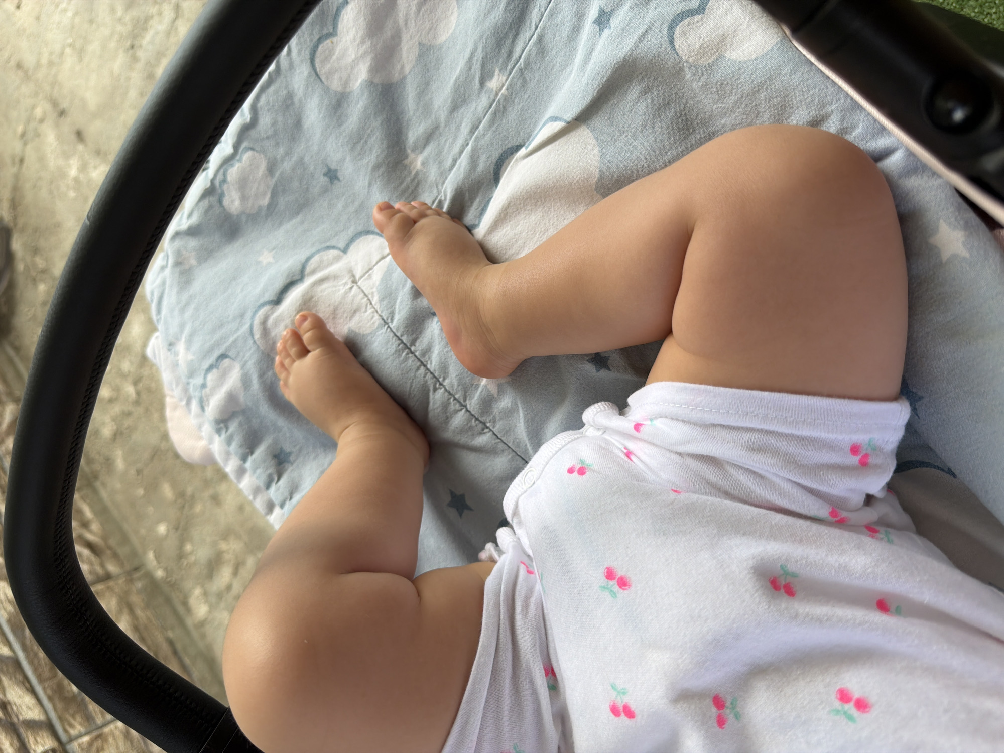 A close-up, high-angle shot shows the legs and feet of a baby lying on a light blue blanket with a cloud and star pattern. The baby is wearing a white onesie with small pink cherry motifs. To the left of the baby's legs, a black handle of a stroller is visible. The background appears to be an outdoor setting, possibly a patio or sidewalk, with a textured surface and a hint of greenery. The image does not contain enough information to determine the specific location within Rusokastro, Bulgaria.