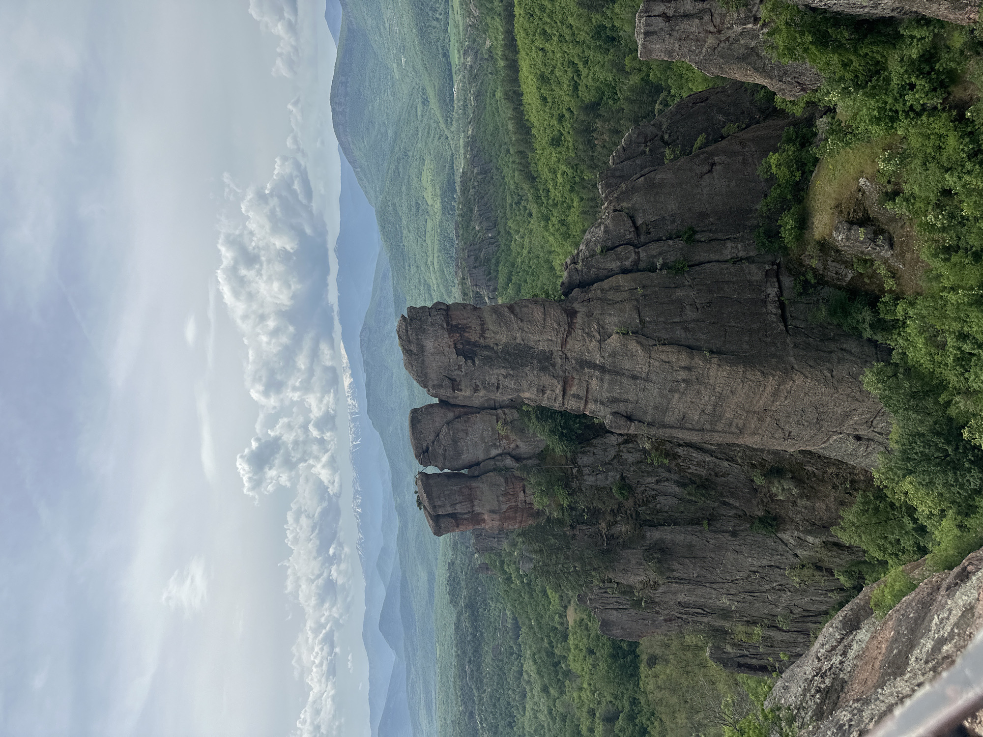 This is a landscape photograph taken in Belogradchik, Bulgaria. In the foreground is a striking rock formation. Beyond the rock formation is a vast expanse of green, forested hills. In the far distance, there are hazy mountain ranges. The sky is partly cloudy, with a mix of blue sky and puffy white clouds. The image gives a sense of height and vastness.