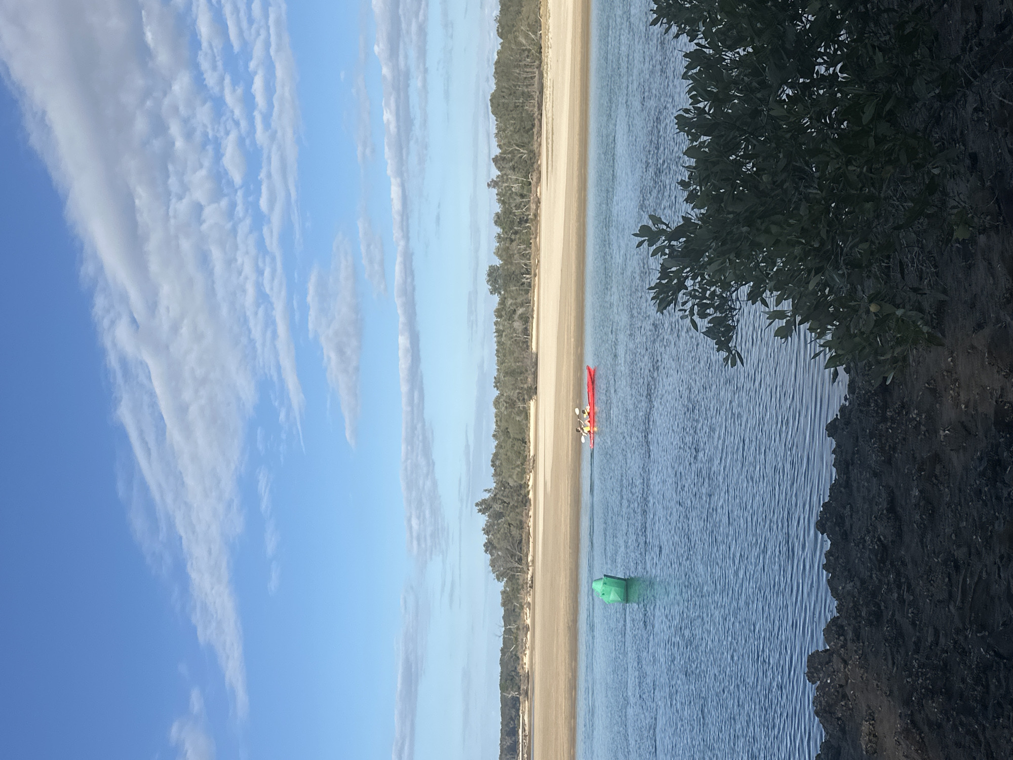 In Nambucca Heads, Australia, a serene scene unfolds along a body of water. The sky above is a vibrant blue, adorned with scattered white clouds, creating a picturesque backdrop. In the mid-ground, a bright red kayak glides through the calm water, carrying two individuals paddling in unison. To the left of the kayak, a green buoy stands tall, marking its presence in the water. Beyond the kayak, a sandy beach stretches across the horizon, backed by a dense line of trees. The foreground is occupied by dark, rocky terrain, partially obscured by lush green foliage.