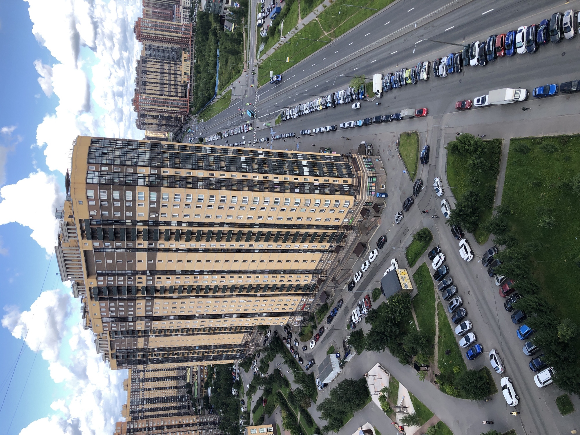 An aerial view captures a cityscape with towering residential buildings and a busy road. The foreground features a modern, multi-story apartment complex with a distinctive tan and brown facade and numerous balconies. Adjacent to this building is a smaller structure, possibly a commercial space or entrance, with a green and white sign. A tree-lined courtyard with some playground equipment is visible to the left, with a few scattered vehicles parked on a winding road.
The main focus shifts to a long, straight road running parallel to the apartment buildings. This road is heavily trafficked, with numerous cars parked on both sides and moving vehicles in multiple lanes. Two blue buses are prominently featured on the road, one moving and one stopped at an intersection. The road is flanked by green spaces, including grass and trees, and more residential buildings can be seen in the background. The sky above is a bright blue with scattered white clouds. The overall scene depicts a dense urban environment characterized by high-rise apartments and active transportation infrastructure.