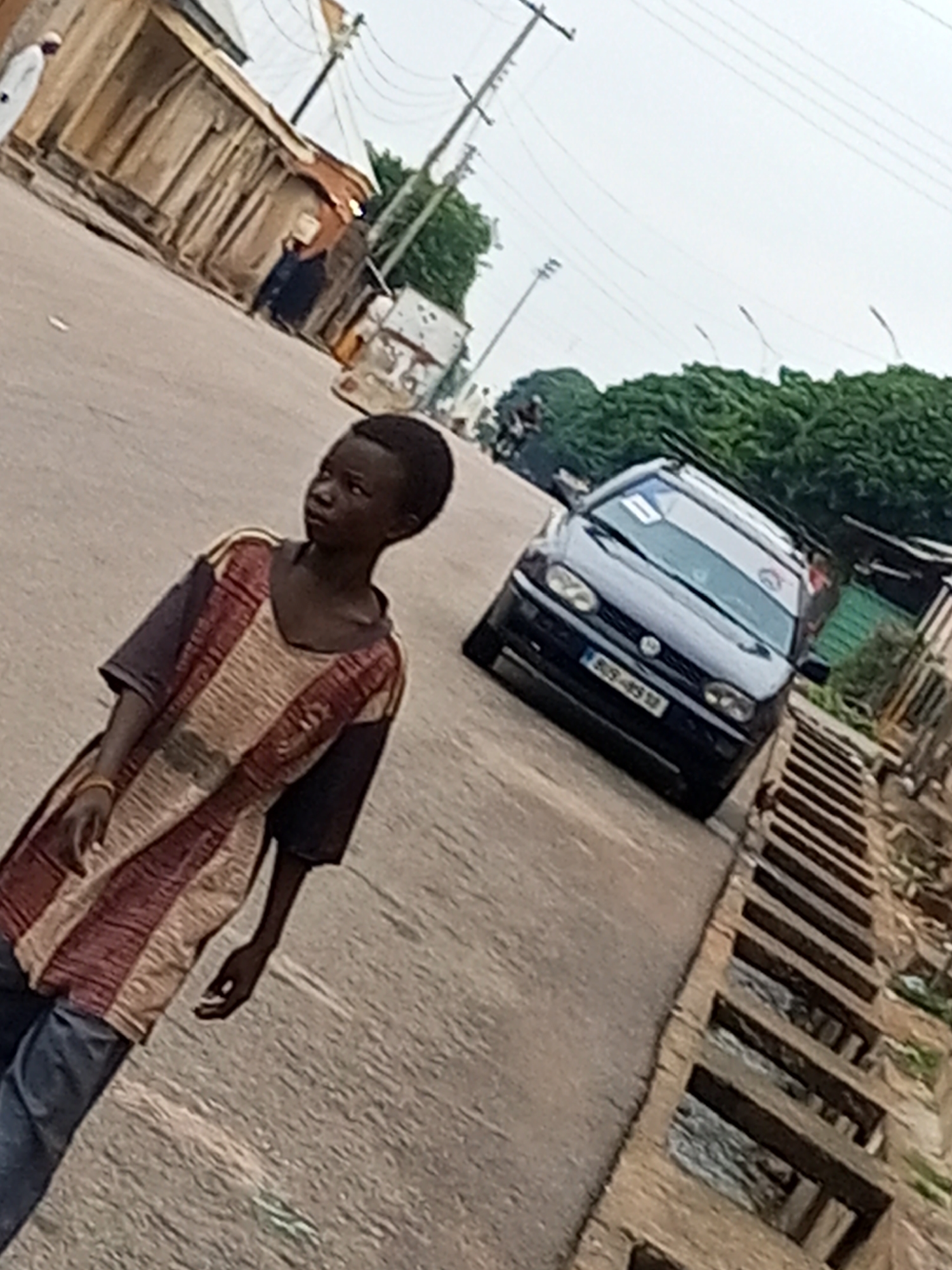 In the city of Arawa, Nigeria, on an overcast day, a young boy of African descent stands on a paved road. He is wearing a vibrantly colored, patterned tunic with horizontal stripes of maroon, beige, and dark blue, and blue jeans. His right arm is extended forward, and he is looking to his left. Behind him, a dark-colored Volkswagen Golf car is parked at an angle on the road, with a roof rack. The car's license plate is visible, reading "B09-4512". To the right of the car, a wooden drainage grate runs along the side of the road, revealing water below. In the background, there are buildings with corrugated metal roofs, trees, and power lines.