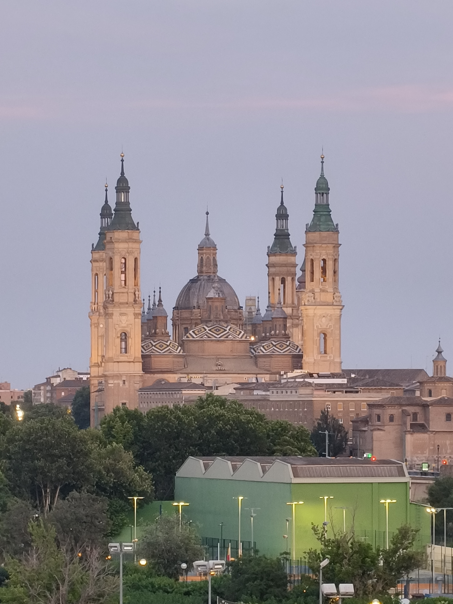 The image shows the Basilica of Our Lady of the Pillar in Zaragoza, Spain. The basilica is a large, ornate building with multiple towers and domes. The towers have green spires and the domes are decorated with patterned tiles. The basilica is surrounded by other buildings, trees, and a modern green structure with street lamps in front. The sky is a soft, pale color, suggesting dawn or dusk.