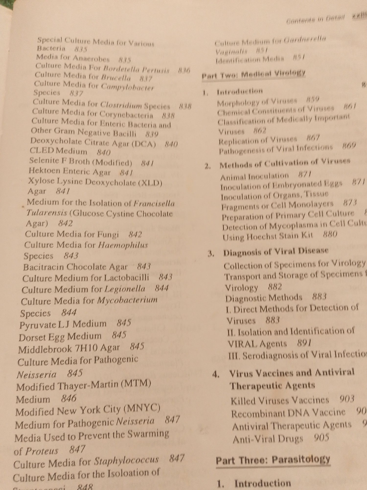 This is an image of a page from a book, likely a textbook or reference guide, focused on medical microbiology and virology.
The subjects are text and the page itself. There are no people, objects, or scenes depicted beyond the printed material. The setting is implied to be a library or study area where someone is reading or referencing this book.
The activity taking place is reading and information retrieval from the book. The notable details are the printed text itself, which lists various culture media, their applications, and page numbers for further information. It also details sections on Medical Virology and Parasitology, including topics like virus introduction, cultivation, diagnosis, vaccines, and therapeutic agents.
Visible text includes headings like "Special Culture Media for Various Bacteria," "Part Two: Medical Virology," "Part Three: Parasitology," and numerous sub-headings and entries listing specific media, diseases, and concepts with corresponding page numbers. There are no time of day, weather, location cues specific to Jalingo, Nigeria, or emotions conveyed by the image.