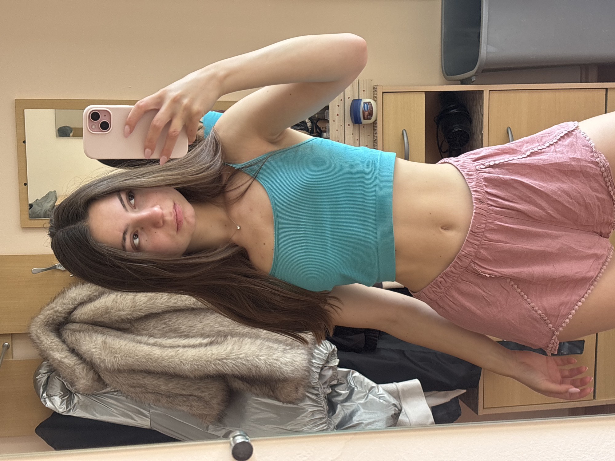 A young woman with long brown hair takes a mirror selfie in what appears to be a bedroom. She is wearing a turquoise crop top and pink pajama shorts. She is holding up a pink phone to take the picture. Behind her, a closet door is open, revealing several coats hanging inside. To her right, a wooden dresser with drawers is visible, along with a small jar of Vaseline on top and a grey trash can. The room is well-lit, suggesting it is daytime. The scene is consistent with a personal living space, and the details of clothing and personal items suggest a relaxed, informal setting. There are no identifiable landmarks or context clues to pinpoint the location to Varna, Bulgaria.