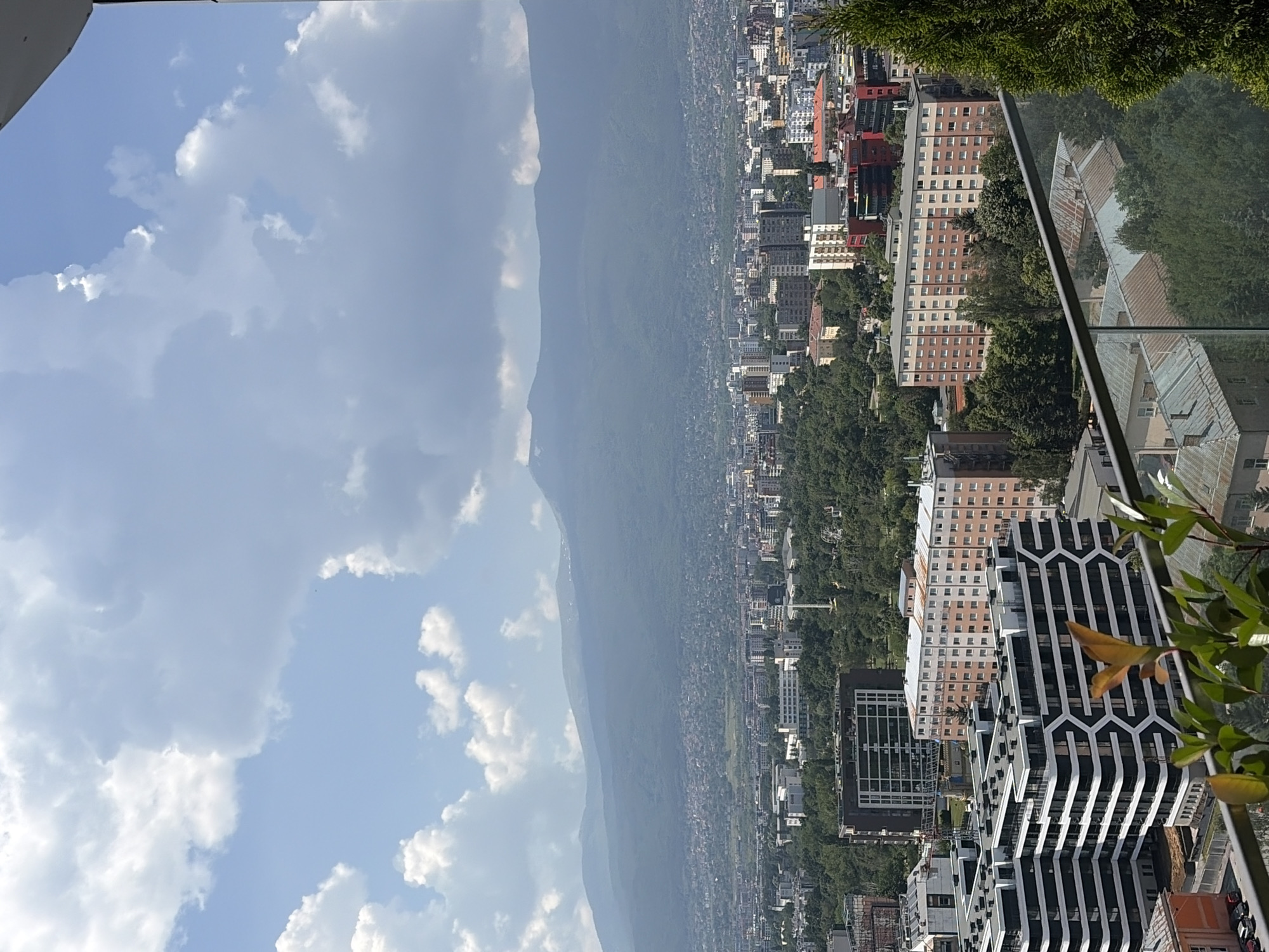 This media file is a vertical, wide-angle, high-angle shot of a cityscape at the base of a large, misty mountain. The sky is a bright blue with scattered white, fluffy clouds. The city is densely packed with buildings of varying heights and architectural styles, suggesting an urban environment. The buildings in the foreground appear modern, with some featuring geometric patterns. Further into the distance, the city stretches towards the mountain, which is covered in lush green trees. The time of day appears to be daytime, with bright sunlight. The weather seems fair. There is no visible text in the image. The overall impression is one of a sprawling city nestled against a natural landscape.