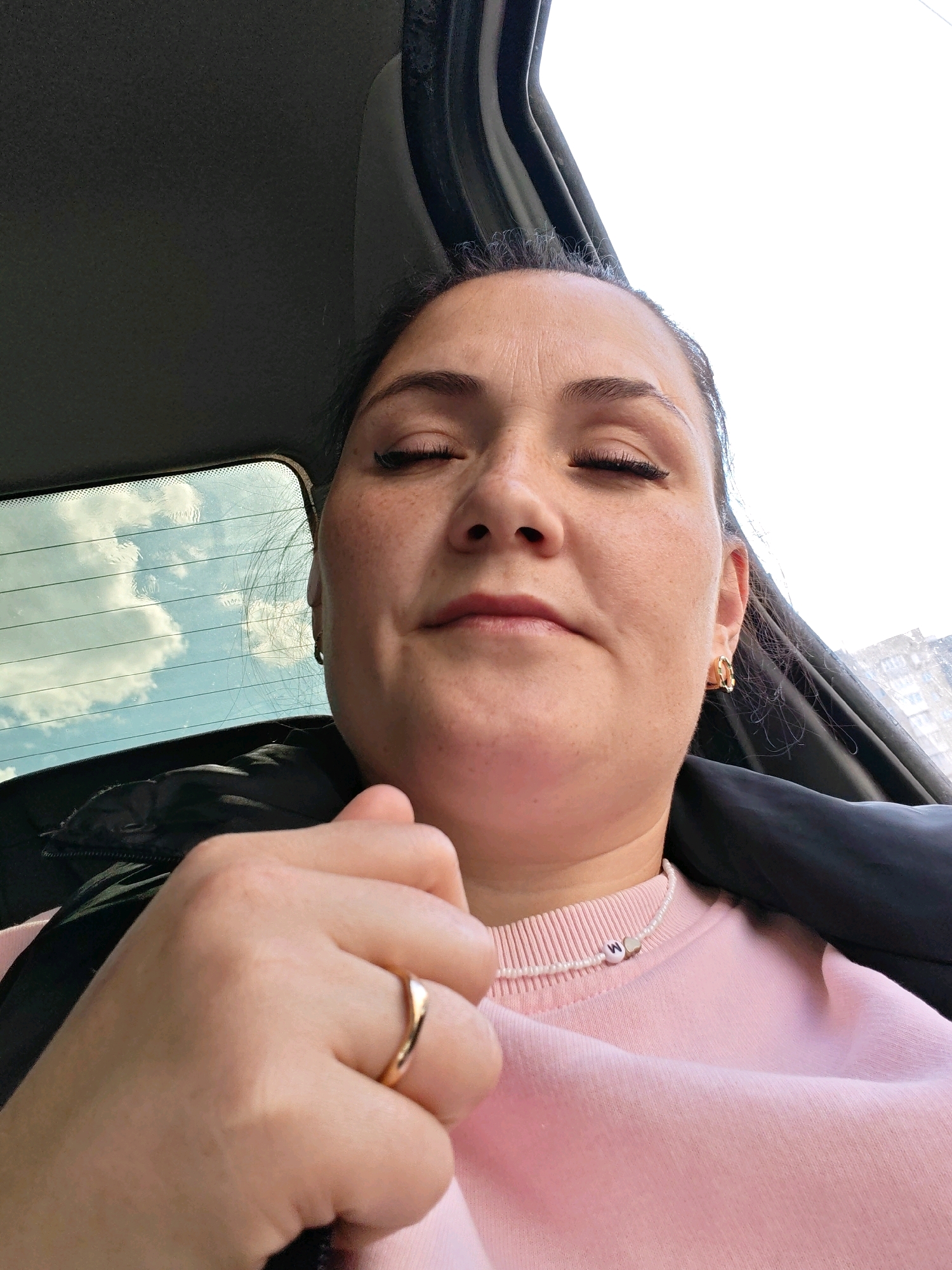 In the original image, a woman is seen from a low-angle perspective, likely taken inside a vehicle. Her eyes are closed, and her head is tilted back slightly. She is wearing a pink sweatshirt with a beaded necklace that has the letter "M" on it. She also has a gold ring on her left hand and gold hoop earrings. The background suggests an urban environment, with a building visible through a car window. The sky is bright, with some clouds. The overall impression is a candid moment, possibly captured while traveling. There are no explicit clues to pinpoint the exact location within Perm, Russia.