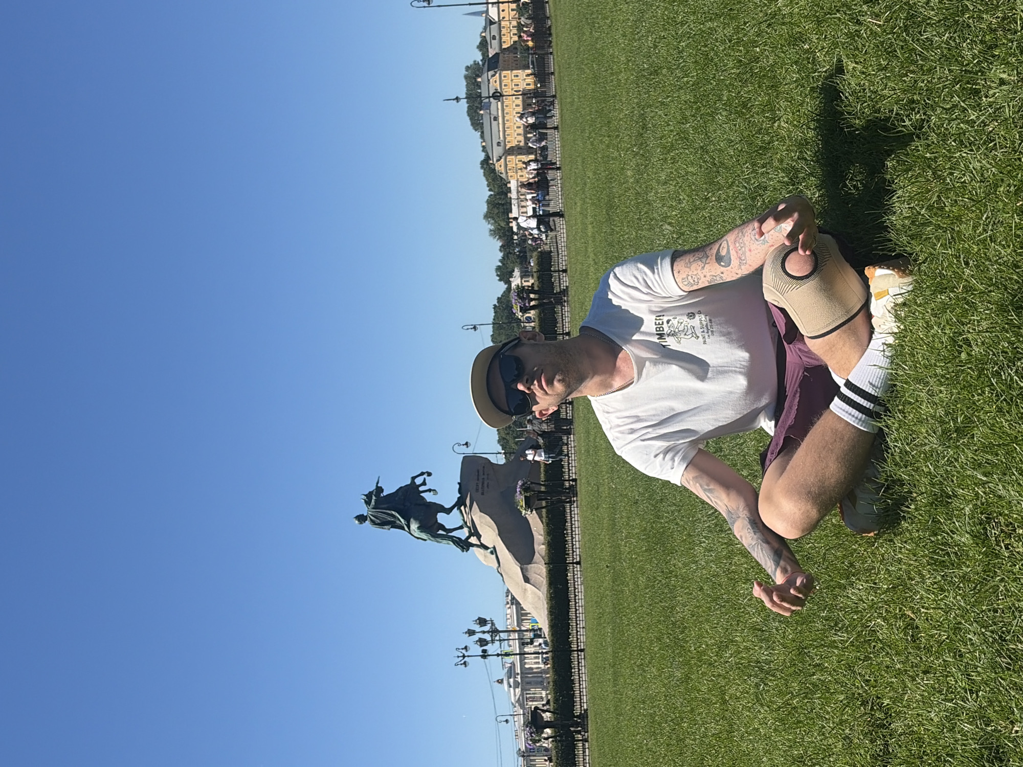A man with tattoos on his arms and wearing a white t-shirt, burgundy shorts, and a baseball cap is meditating in a cross-legged position on a green lawn. In the background, a large equestrian statue of Peter the Great stands on a granite pedestal. Behind the statue, several neoclassical buildings with yellow facades are visible, characteristic of Saint Petersburg, Russia. The sky is clear and blue, indicating a sunny day.