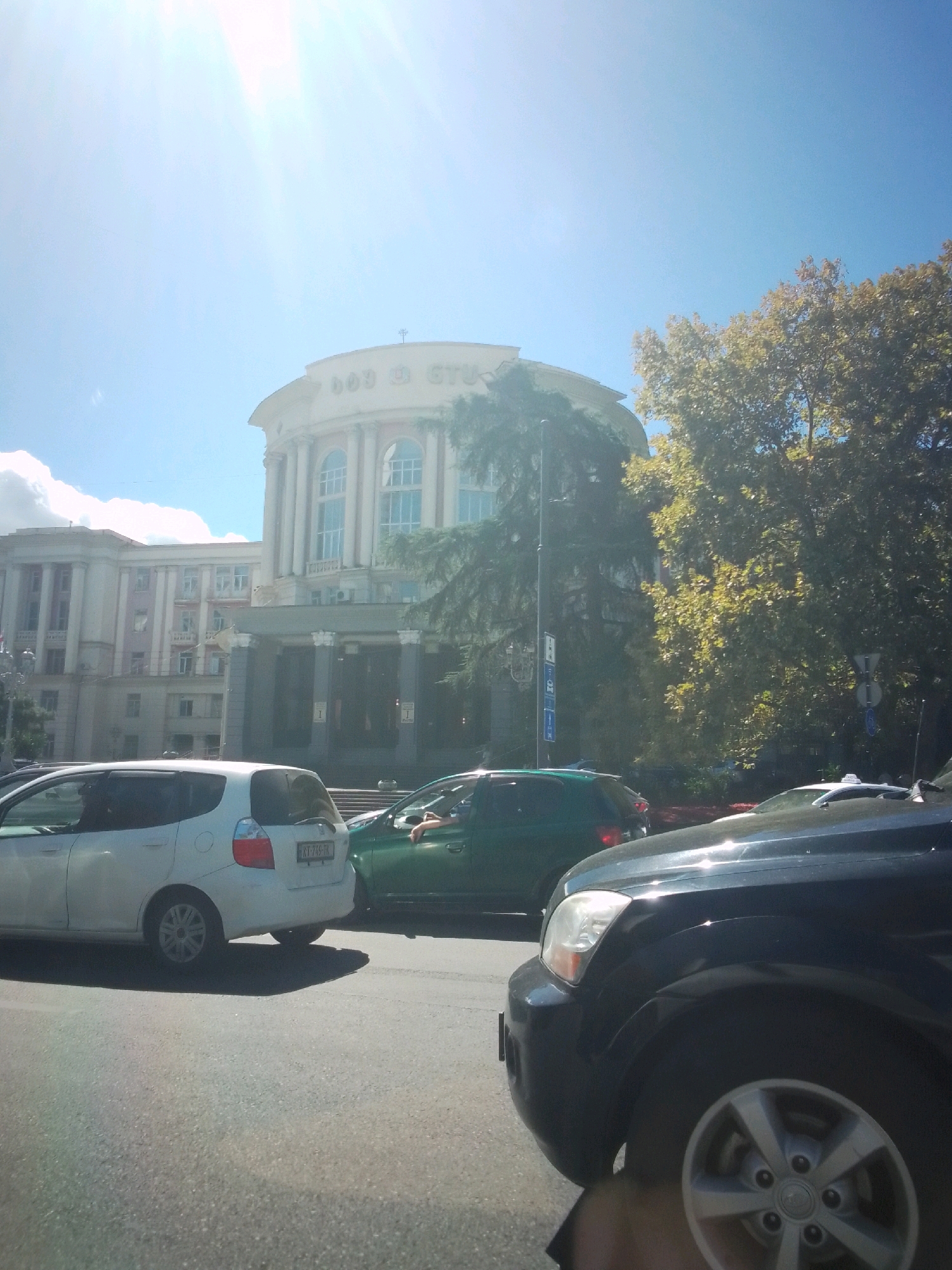 This image depicts a street scene in Tbilisi, Georgia, with a focus on the facade of a large, circular building topped with a cross. The building appears to be a public institution, possibly educational, with large arched windows and classical architectural elements such as columns. The name "BOB U. STU" is partially visible on the building's facade. In the foreground, several cars are visible, including a white hatchback and a dark SUV, suggesting a moderate level of traffic. A green SUV is also present, with a person's arm visible resting on the open window. Lush trees with yellowing leaves, indicative of autumn, flank the right side of the building and partially obscure the sky. The sky is bright blue with some scattered clouds and strong sun glare, creating lens flares in the upper left quadrant of the image. The overall atmosphere is that of a sunny day in an urban setting.