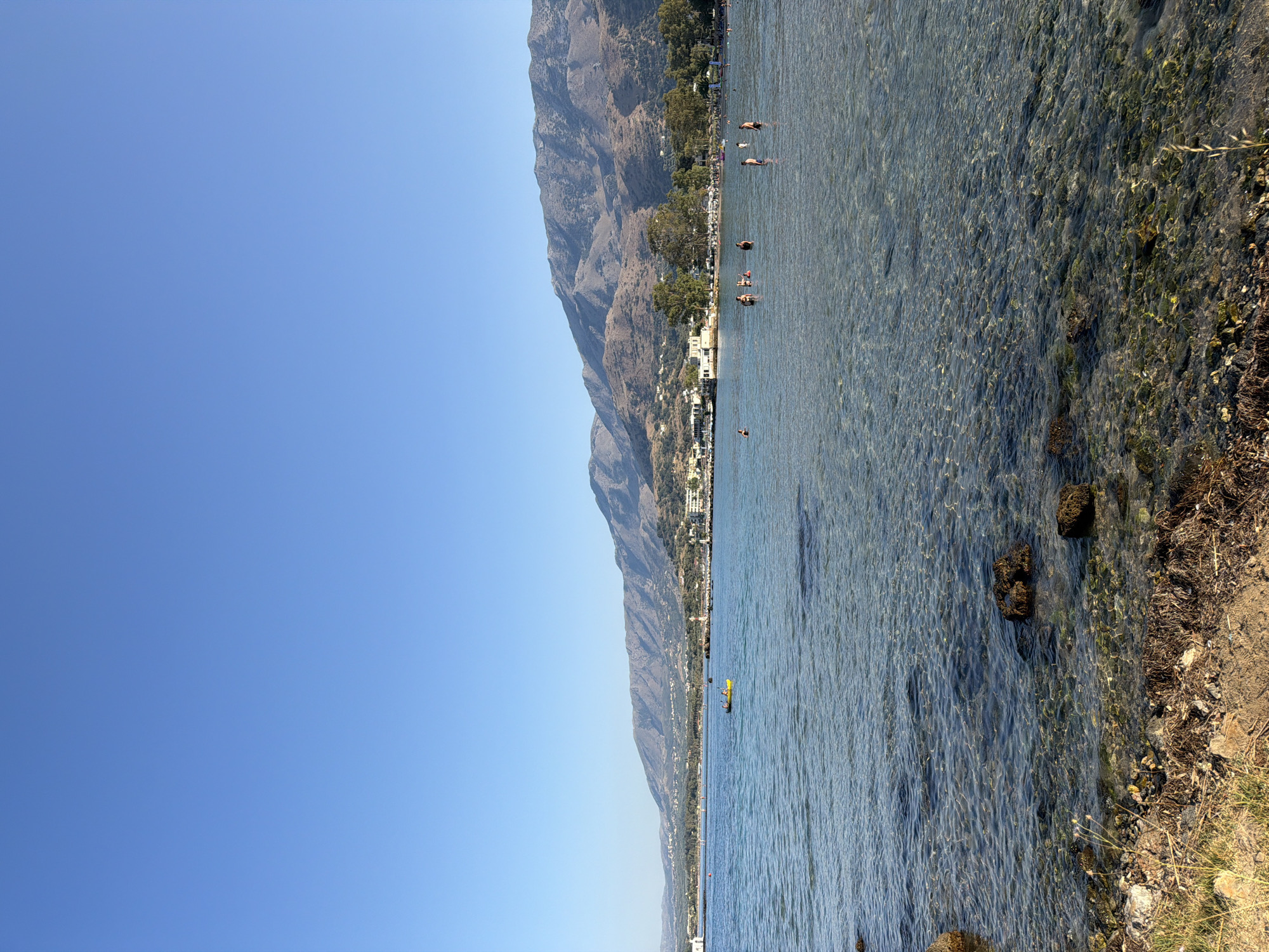 This is a wide-angle, eye-level, outdoor, daytime photograph of a scenic coastal view in Georgioupoli, Greece. The foreground features a rocky, sandy shoreline with dry grass and seaweed. The clear, shallow water of the sea extends from the shore, with sunlight reflecting off the seabed and illuminating submerged rocks and vegetation. In the middle ground, several people are visible wading and swimming in the water. A yellow kayak with two occupants is also present in the water. In the background, a long stretch of coastline is visible, with buildings, trees, and a prominent mountain range under a clear blue sky. The overall scene depicts a tranquil summer day at the beach.