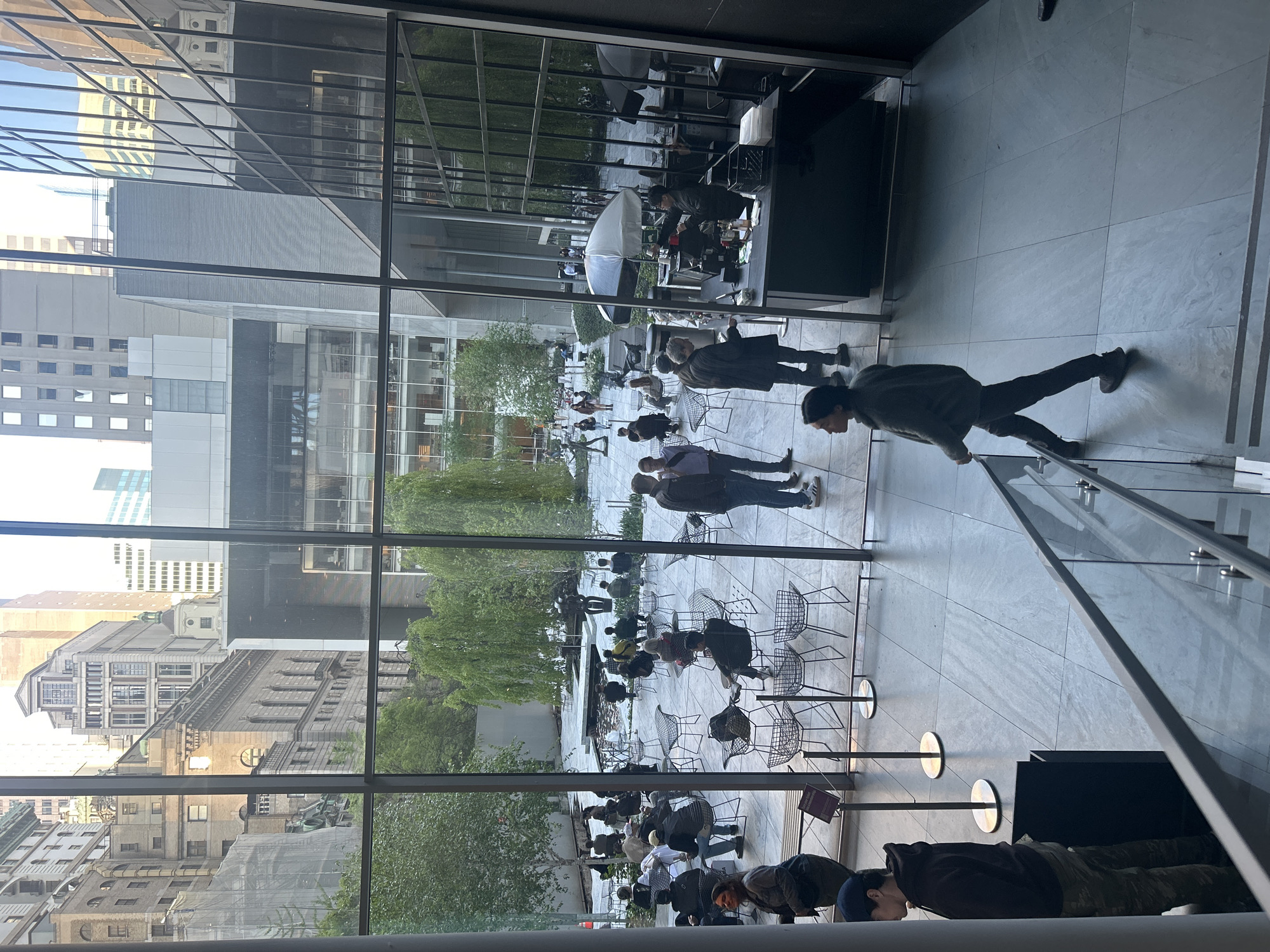 The image captures an indoor scene looking out onto an outdoor courtyard. Through a large glass window of what appears to be an interior space, a courtyard filled with people sitting in chairs can be seen. Trees and shrubbery are scattered throughout the courtyard, providing a natural, green contrast to the urban environment. The reflections in the glass indicate a cityscape beyond the courtyard, characteristic of New York City. A person is walking on a light-colored tile floor in the foreground. Metal stanchions with circular bases are also visible in the foreground. The overall atmosphere is one of urban tranquility, where people are enjoying an outdoor space within the city.