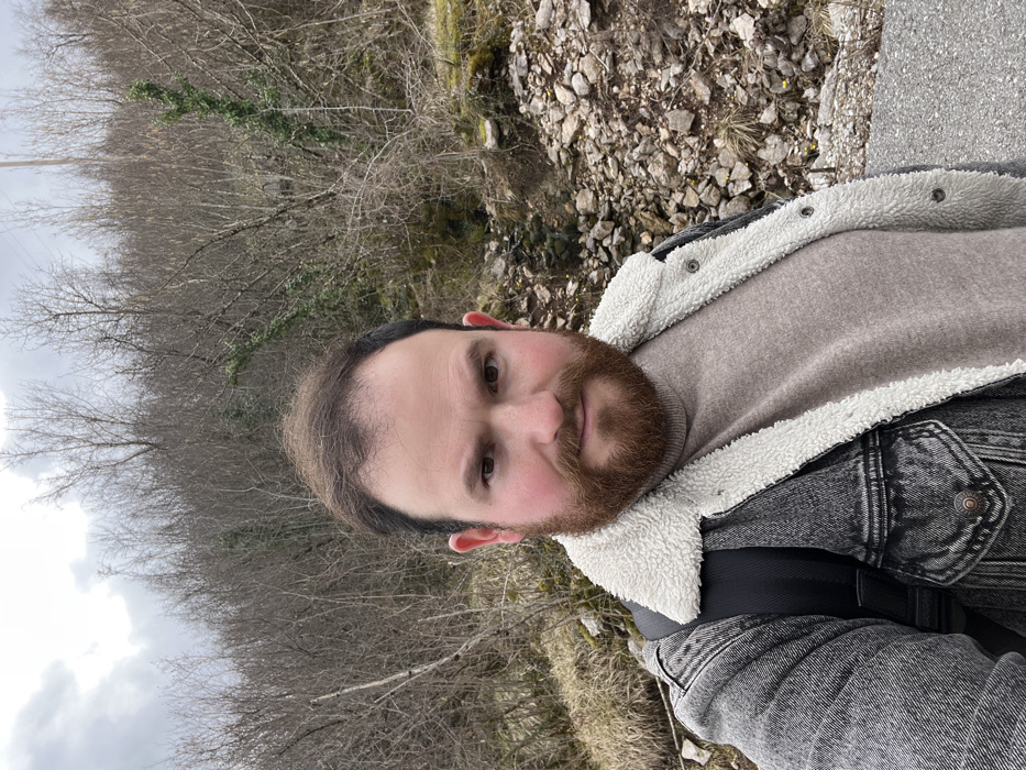 An adult male is depicted in a close-up, chest-up shot against a natural, rocky hillside environment. The scene is located in Vrela, Montenegro. The man, with a reddish-brown beard and dark hair, stares directly at the camera with a neutral expression. He wears a grey acid-washed denim jacket featuring a white shearling-like collar and lining, over a light brown turtleneck sweater. A black backpack strap is visible over his left shoulder.
The background consists of a steeply sloped terrain covered with dense, leafless deciduous trees and shrubs, suggesting a cooler season. Patches of dry grass and abundant scattered rocks are visible on the ground of the slope, particularly on the right side. A narrow, rocky depression, possibly a dry gully or drainage, runs diagonally through the mid-ground. Parts of a utility pole are discernible among the bare branches in the upper left background. The sky is uniformly overcast and grey. The subject appears to be standing on a paved surface, likely a road or path, which is partially visible in the lower right foreground.