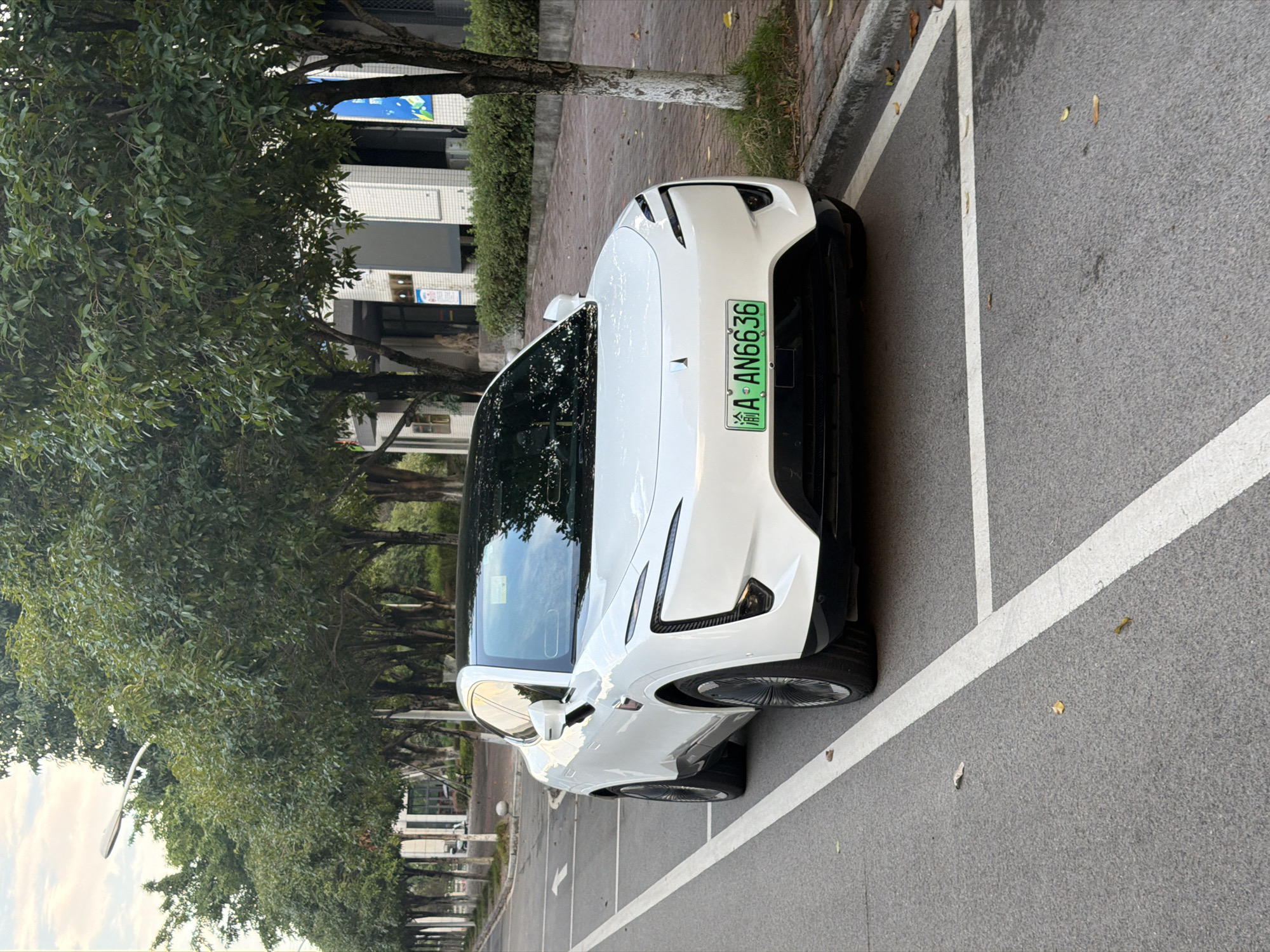 A white electric vehicle, identified by its license plate "渝A AN6636", is parked in a marked parking space on a paved road. The car is positioned facing forward, with its front end visible. The vehicle has a sleek, modern design with distinctive headlights. The road is lined with trees, and a sidewalk with brick paving is visible to the right of the road. In the background, there is a building with a tiled facade and some signage. The sky is overcast. The scene appears to be in the city of Jiangjin, China.