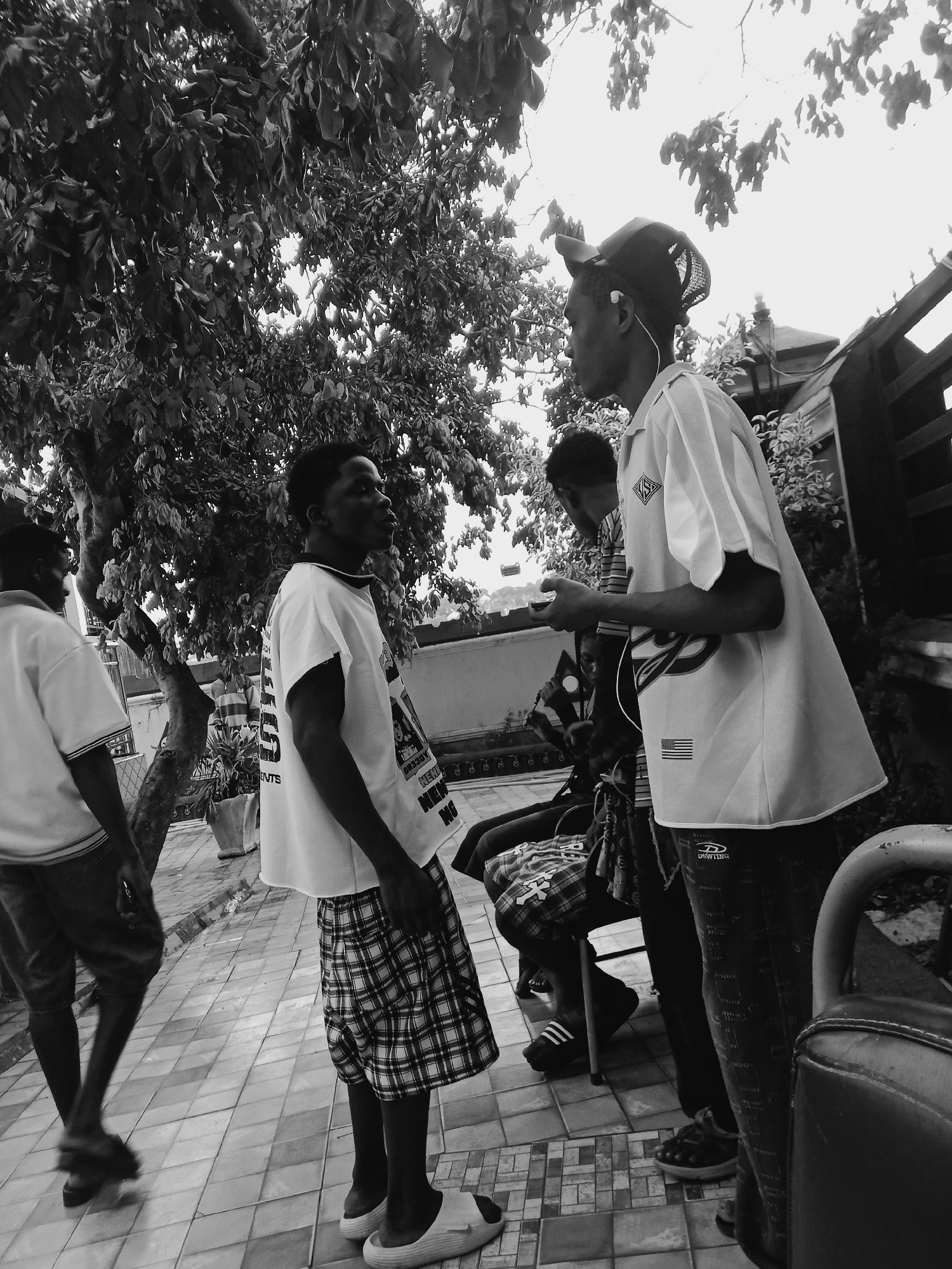Onitsha, Nigeria: Four young men are in an outdoor scene. One man walks away, wearing shorts and a t-shirt. Another man, wearing plaid shorts and a t-shirt, stands talking with a third man, who is wearing a cap and an American-flagged shirt with an earbud in his ear. The third man appears to be looking at a device in his hand. A fourth man sits on a bench. Trees and foliage surround the scene, and a building is visible in the background.