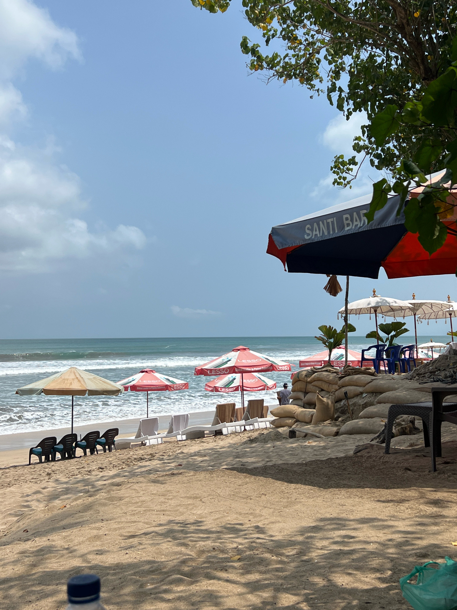 The image depicts a beach scene in Kuta, Indonesia, likely on a sunny day with scattered clouds. In the foreground, a sandy beach is visible, with shadows cast by overhead foliage. A partially filled plastic water bottle with a blue cap is in the lower portion of the frame. Further back, several lounge chairs are arranged on the sand, some with blue cushions. Beyond the chairs, beach umbrellas are set up. Some umbrellas are red and white with the word "LESSO" printed on them, while one is a faded beige. A large umbrella with "SANTI BAR" printed on it in white letters against a dark background is prominently displayed on the right side, partially obscuring some of the other umbrellas. In the middle ground, sandbags are piled up, creating a raised barrier. Behind the sandbags, several white umbrellas with decorative finials are arranged, and blue plastic chairs are visible. The ocean with moderate waves is in the background, stretching towards the horizon under a pale blue sky. A lone figure, appearing to be a person, is seen standing near the water's edge. The overall impression is of a beach resort area, with amenities for visitors.