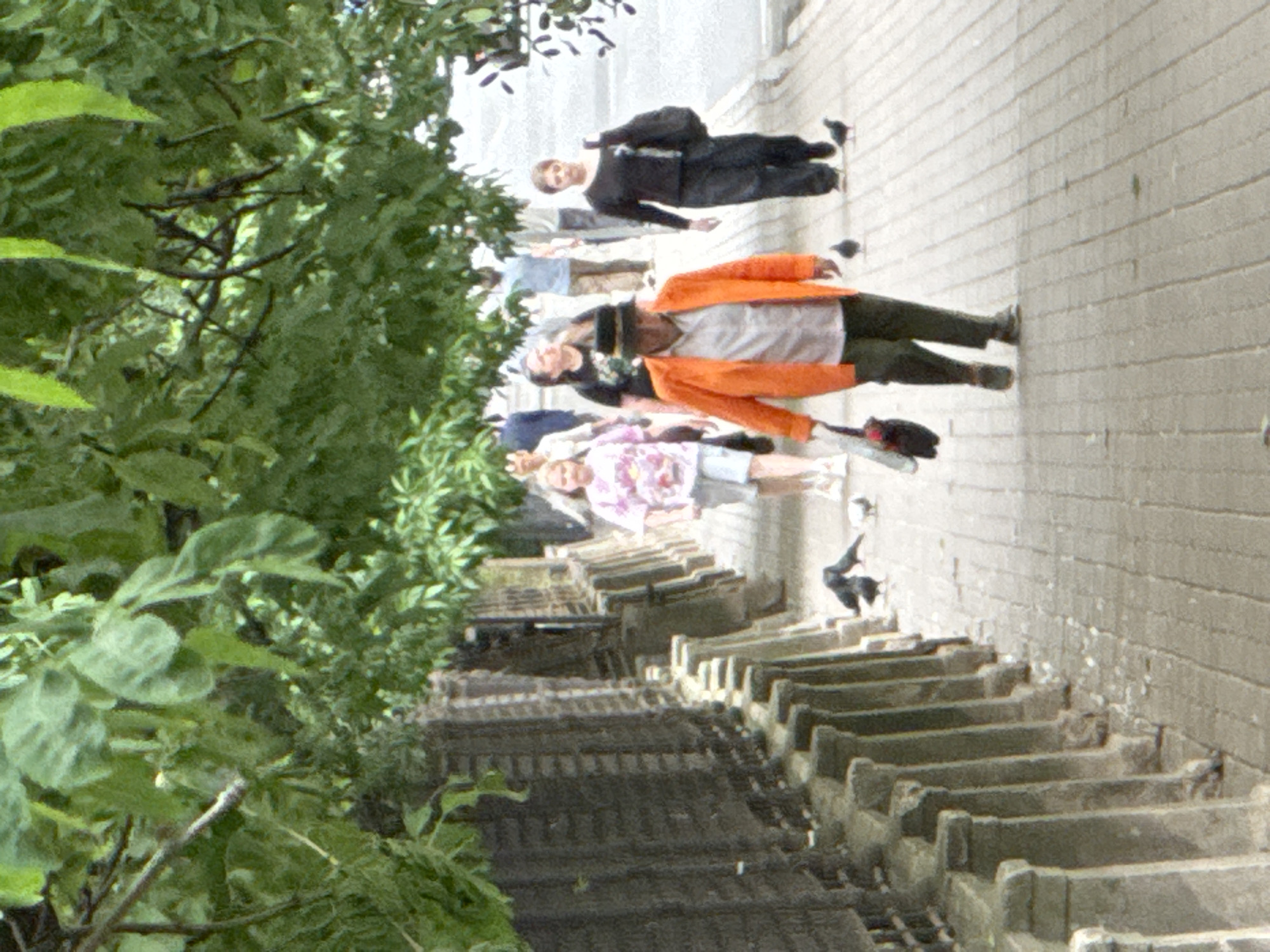 This is a medium shot of people walking on a sidewalk in Saint Petersburg, Russia. The sidewalk is paved with light-colored bricks and bordered by a dark, textured wall on the left. Trees with green leaves hang over the sidewalk, casting shadows.
In the foreground, a person wearing an orange jacket, a white shirt, dark pants, and a hat is walking away from the camera. They are carrying a black bag in their left hand. Two pigeons are walking on the sidewalk near them.
Further down the sidewalk, a person in dark clothing and carrying a black bag is walking towards the camera. Behind them, a girl in a pink patterned shirt and shorts is walking away from the camera. Other figures are visible further in the background, including a man in a blue shirt and a woman with dark hair. The overall atmosphere is casual and urban.