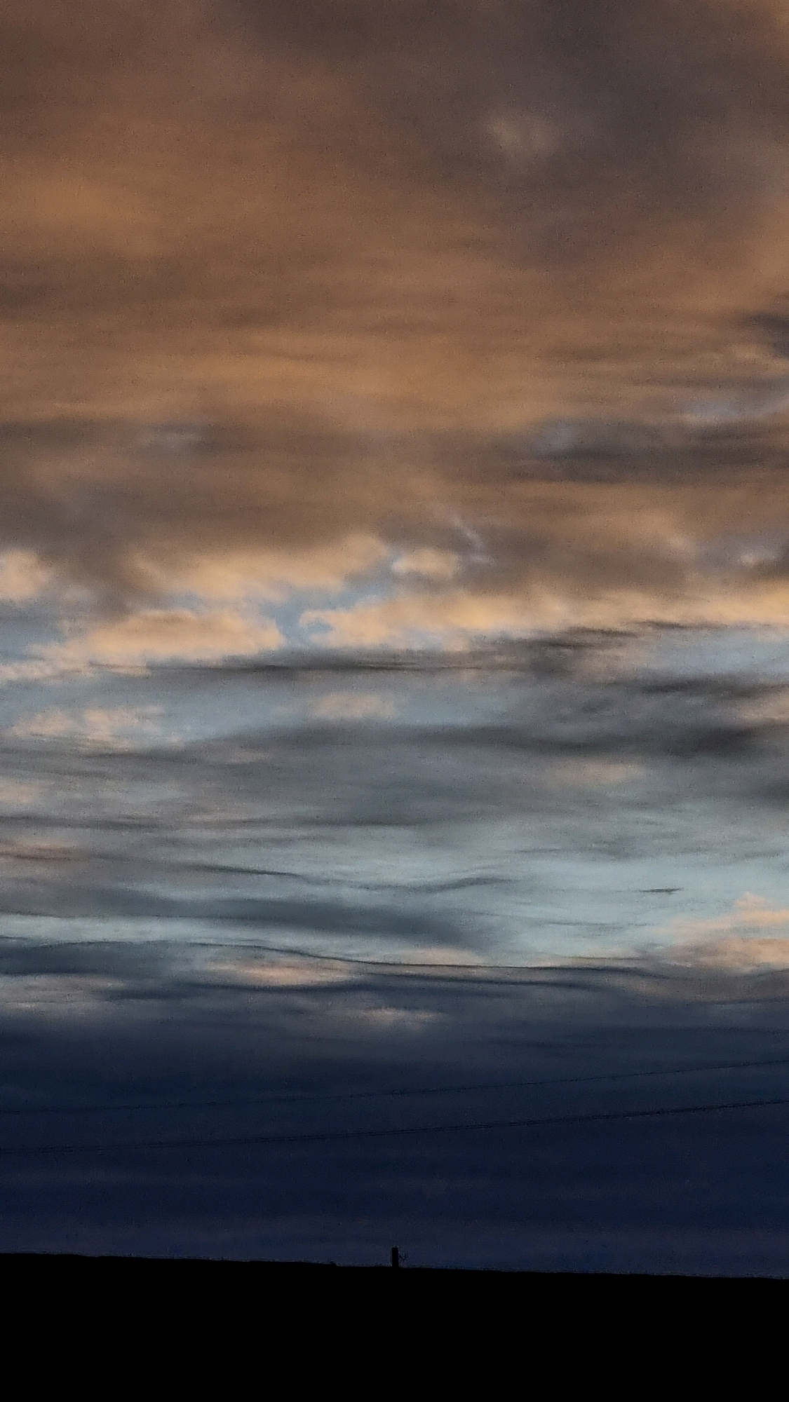 This is a photograph of a cloudy sky over a silhouetted landscape. The clouds are varied in color, with streaks of brown, grey, and blue, suggesting a sunset or sunrise. The bottom of the image is a dark, flat silhouette of the horizon, with a single vertical post visible in the distance. There are no people or specific landmarks that can identify the location as Vysokoe, Russia. The image captures a natural scene with a focus on the atmospheric conditions of the sky.