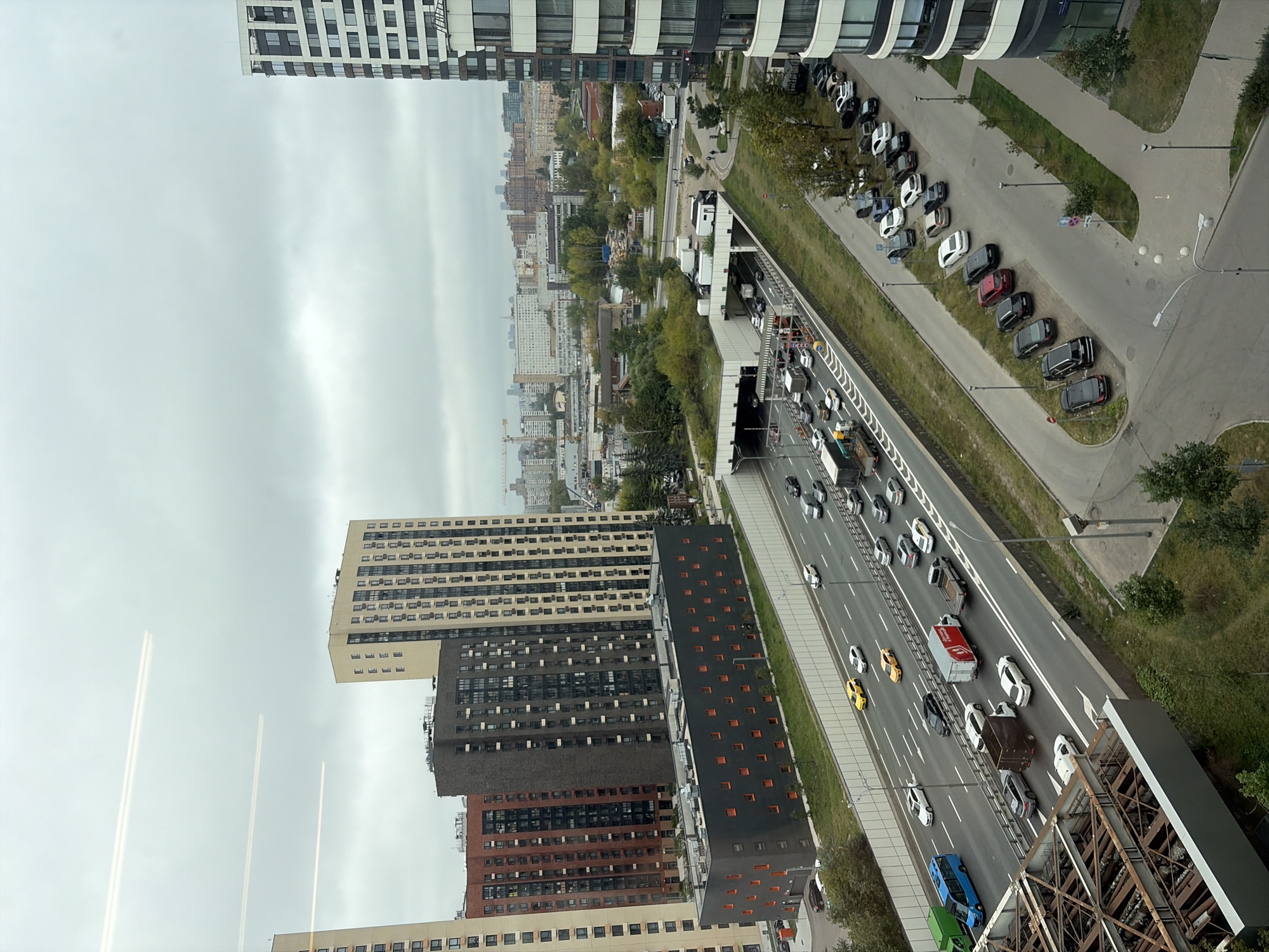 The image shows an aerial view of a highway passing through an urban area with multiple high-rise buildings in Moscow, Russia. The highway is a multi-lane road with significant traffic. Several cars, trucks, and a blue bus are visible on the highway. There is a tunnel entrance on the left side of the highway, covered with a green roof. Several apartment buildings of varying heights and architectural styles are present in the background, including modern high-rises and older structures. The sky is overcast, suggesting a cloudy day. The foreground shows part of a structure with exposed metal beams and concrete. A parking lot with several cars is visible to the right of the highway. The overall scene depicts a bustling urban environment with a focus on transportation infrastructure and residential buildings.