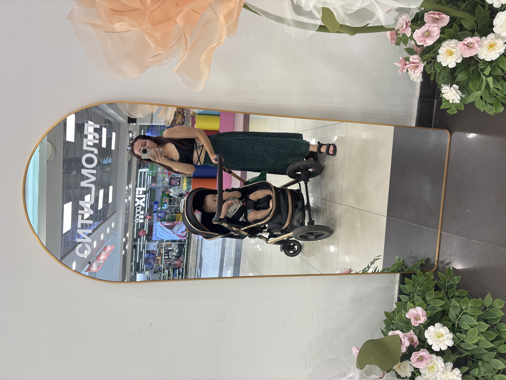 In Naberezhnye Chelny, Russia, a full-length mirror with a gold arch frame is positioned against a white wall. The mirror reflects a scene inside a commercial space, likely a shopping center, with brightly lit ceiling panels and various retail displays visible.
The reflection shows a young woman taking a selfie. She is wearing a black crop top with criss-cross detailing and a long, dark green polka-dot skirt. She is holding a smartphone in her right hand.
To her left, also reflected in the mirror, is a baby in a black and gold stroller. The baby is sitting upright and appears to be looking to the side.
In the foreground, on either side of the mirror, are artificial floral arrangements. The arrangement to the left features green foliage with clusters of pink and white flowers. The arrangement to the right is larger, with translucent peach and white petals, and also includes green foliage. The floor appears to be tiled with a light color in the reflected area, and darker tiles around the base of the mirror.