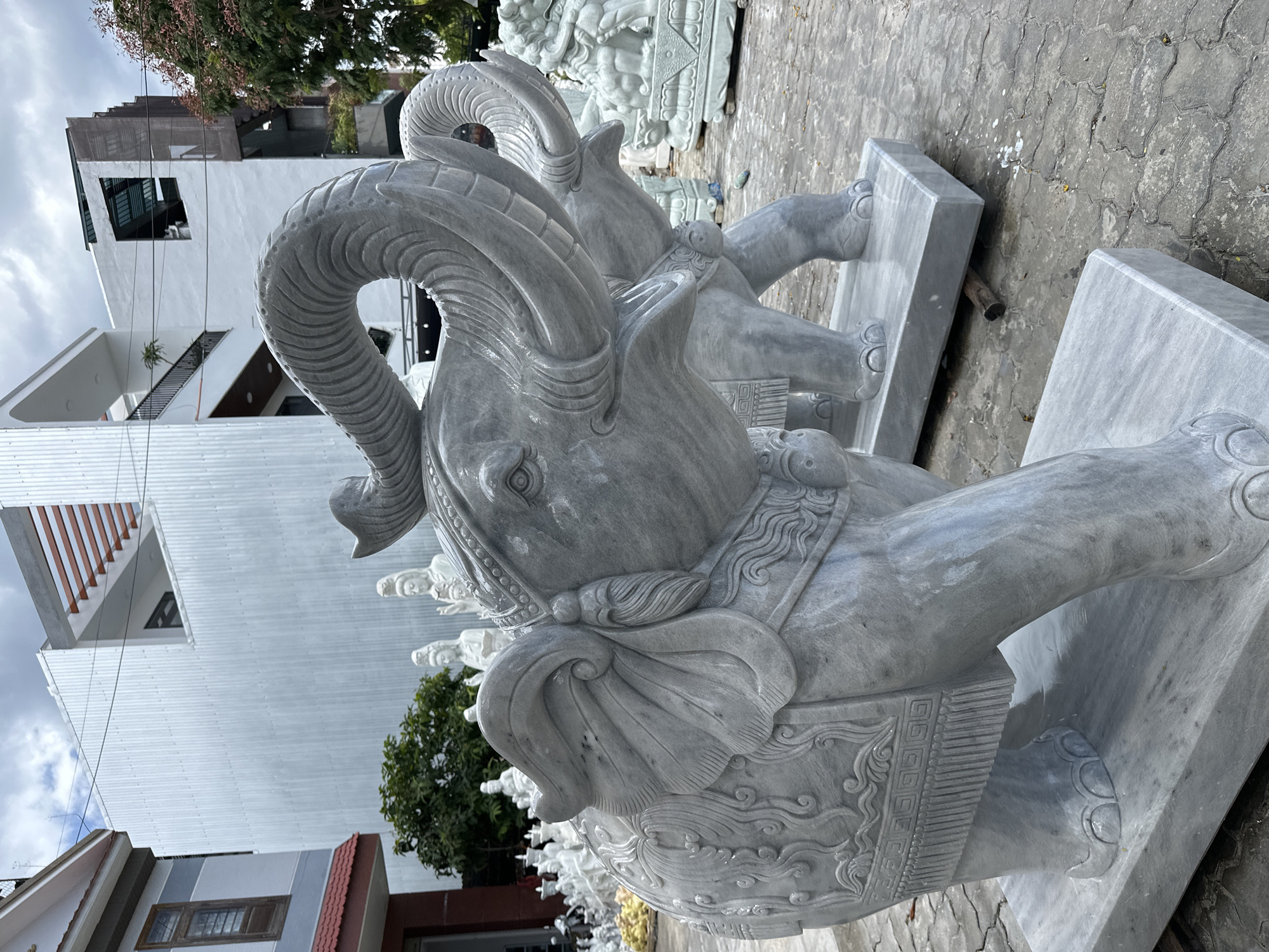 The image shows a close-up view of a large, grey marble sculpture of an elephant in an outdoor courtyard in Da Nang, Vietnam. The elephant sculpture is highly detailed, with intricate carvings on its body and trunk raised upwards. In the background, other marble sculptures of figures, including what appear to be Buddhist deities, are visible, along with modern buildings and trees. The ground is paved with interlocking concrete tiles, and there is a slight sheen on the sculptures, suggesting they may have been recently cleaned or are naturally wet. The overall scene suggests a sculpture workshop or an outdoor display area for stone carvings.