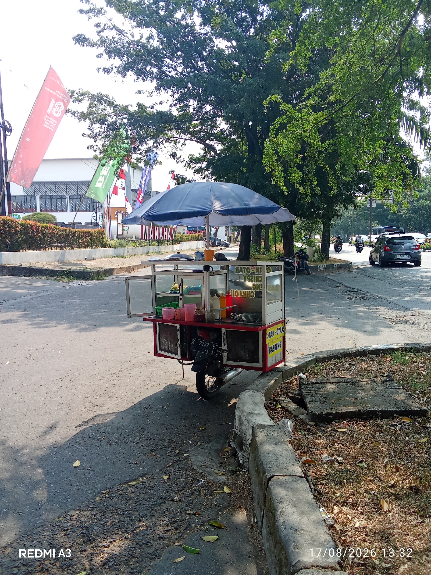 A food cart with a blue umbrella is parked on the side of a road in Surakarta, Indonesia. The cart is attached to a motorcycle and contains food items and ingredients in plastic containers. A sign on the cart reads "MANAHAN", indicating its location. In the background, there are trees, buildings, and other vehicles on the road. The weather appears to be sunny.