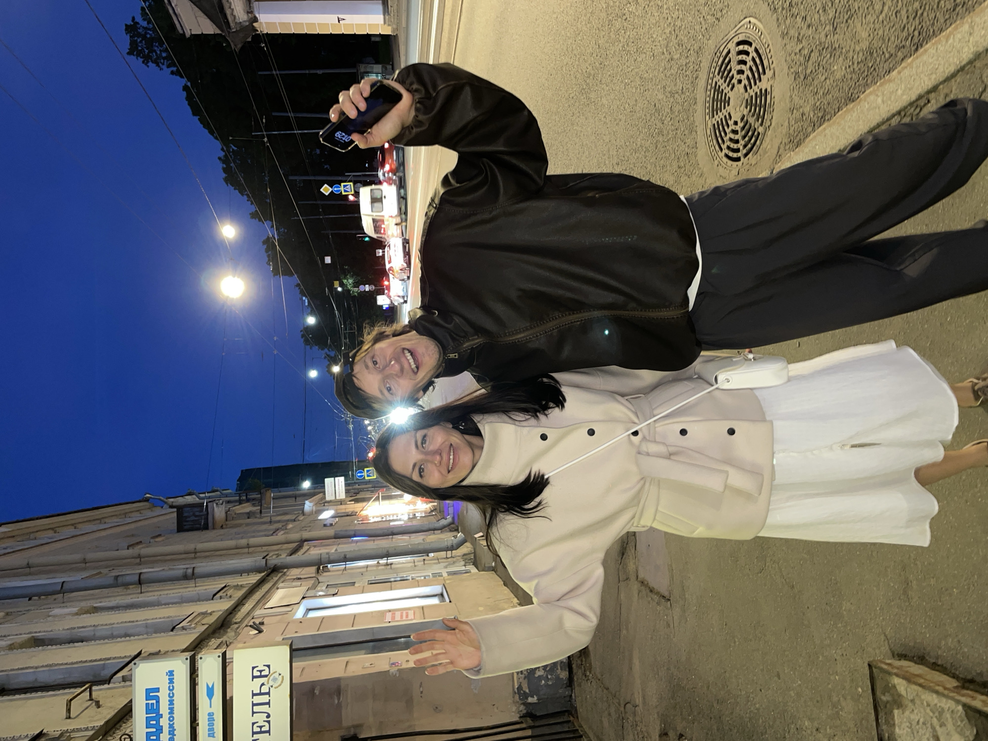 Two individuals, a woman and a man, are standing on a sidewalk in front of a building at night. The woman is on the left, wearing a light-colored coat and a white dress, and is waving with her left hand. The man is on the right, wearing a dark jacket and dark pants, and is holding up a smartphone in his right hand. They are both smiling and looking at the camera. The background shows a street with passing cars, streetlights, and a building facade. The image appears to be taken in an urban environment during dusk or early night.