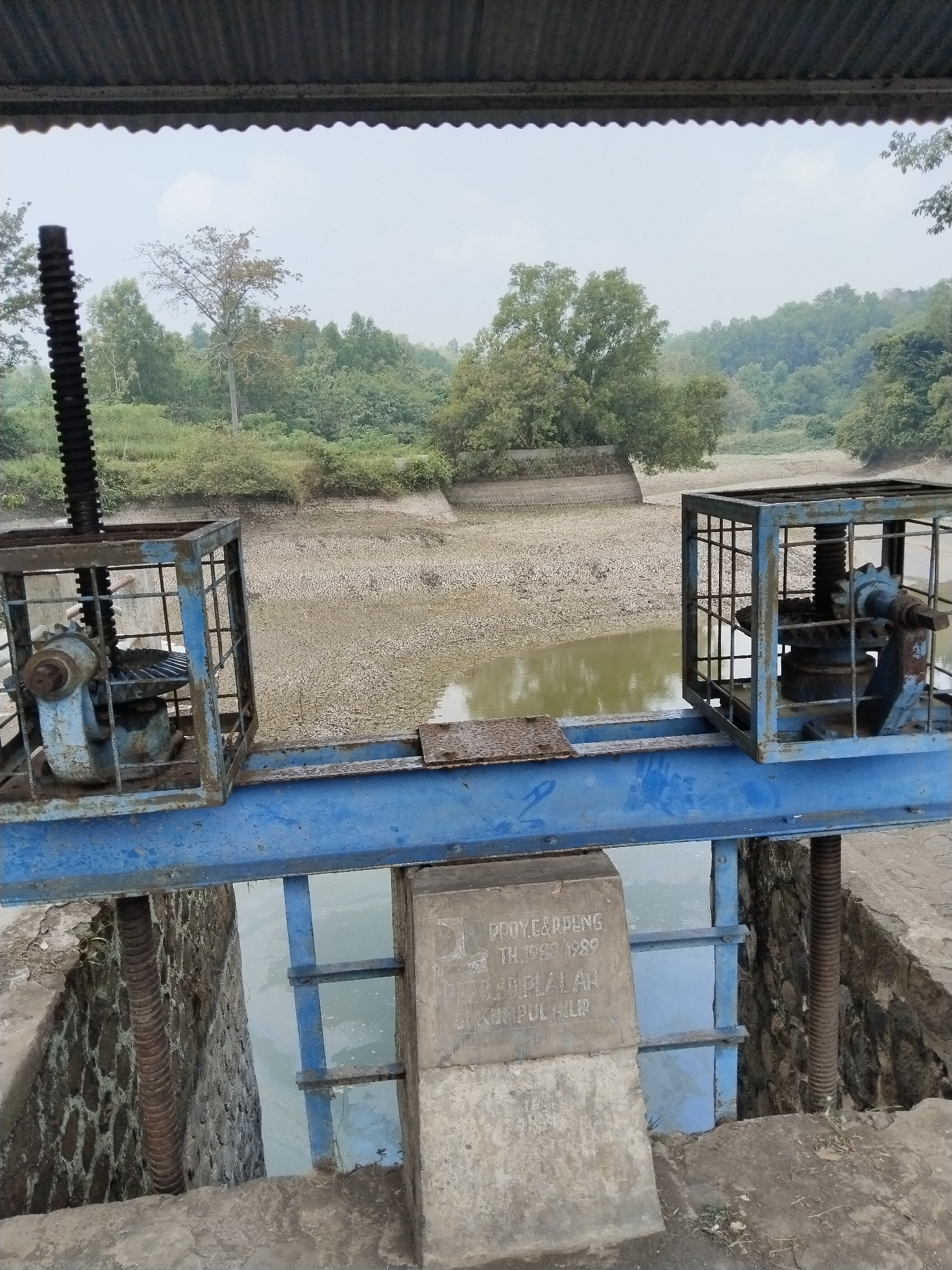 The image displays a water control structure, likely a sluice gate or weir, located in Kumpul Hilir, Unknown, Unknown city. A dark gray corrugated metal roof partially obstructs the top portion of the view. Below, a heavy blue metal beam spans horizontally, supporting two rusted, caged mechanisms. Each mechanism contains exposed gears and a vertical threaded lead screw designed for raising or lowering a gate. The lead screw on the left extends prominently upwards.
Below the main blue beam, a blue metal gate is partially submerged in a narrow channel of murky, greenish-brown water. The body of water beyond this gate recedes into a wide expanse of parched, light brown, cracked earth, indicating a severe drought or extremely low water levels. Concrete or stone retaining walls are visible along the edges of the dry riverbed, leading towards distant rolling hills densely covered in green vegetation. The sky above is overcast and pale gray.
In the lower right foreground, a concrete pillar supports the structure and bears an etched inscription: "PROYEK PPNG TH. 1988/1989 DIR. BB. PALAR KUMPUL HILIR". Threaded metal rods are visible along the stone abutments on either side of the water channel.