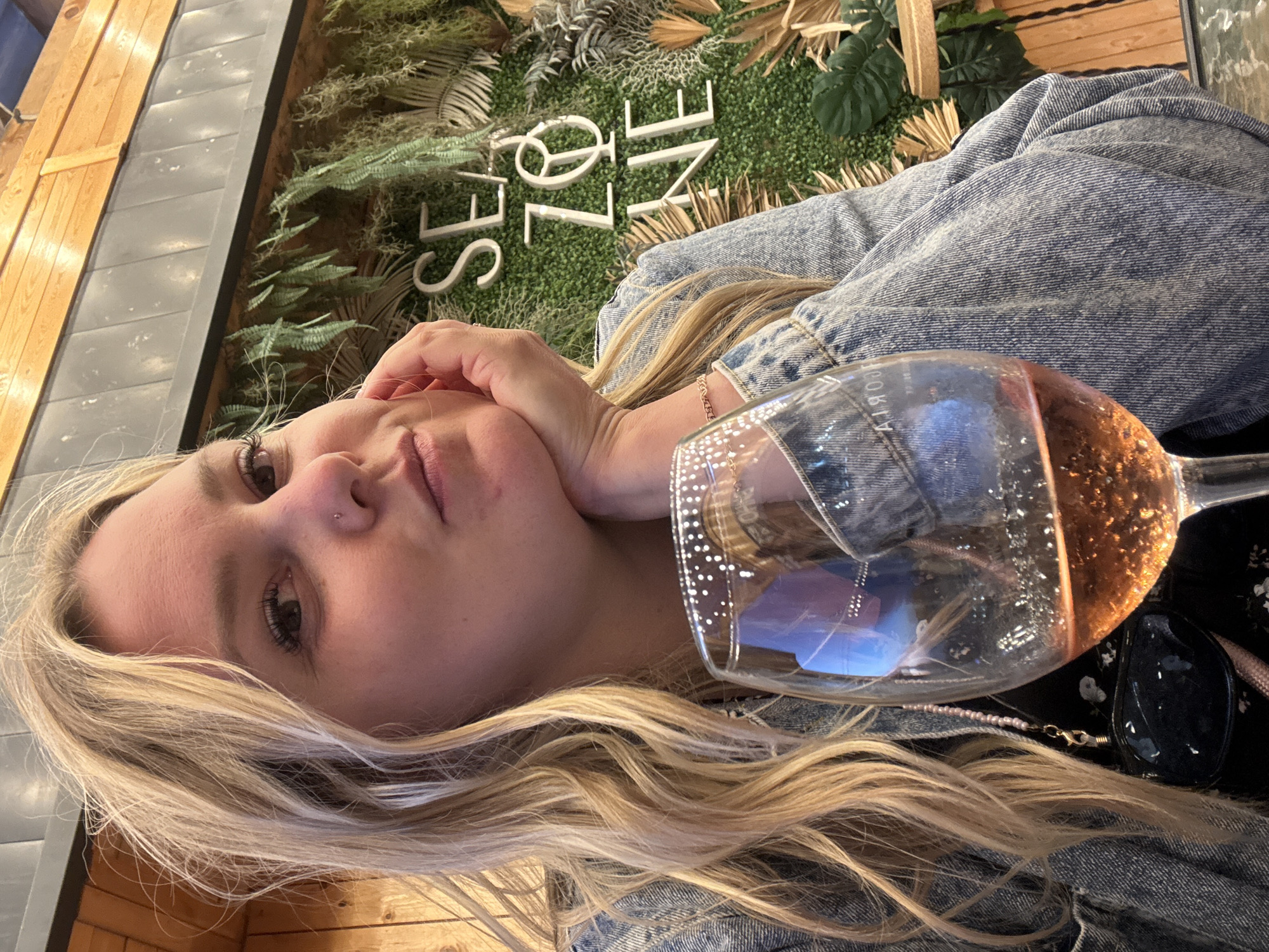 A young blonde woman is captured in a close-up, rotated shot, appearing to be a selfie. She has long, wavy hair, and a small stud nose piercing. Her left hand rests gently against her chin, and she looks towards the camera with a neutral expression. She is wearing a light-wash denim jacket.
In the foreground, a large wine glass filled with a rosé-colored beverage is prominent, partially obscuring her body. Reflections, possibly of the surroundings or a label, are visible within the glass. Nearby, a pair of black sunglasses and a pink beaded chain or necklace are also visible.
The background features a decorative wall adorned with artificial green foliage and various faux plants like ferns and dried palm fronds. On this green wall, large white three-dimensional letters spell out "SEA ZNNE," likely indicating "SEA ZONE" as the name or theme of the establishment. To the left, a wooden paneling structure and a gray slatted wall are visible. The setting suggests an indoor or semi-outdoor space, possibly a restaurant, cafe, or bar, in Sennoi, Russia, where the woman is seen relaxing with a drink.