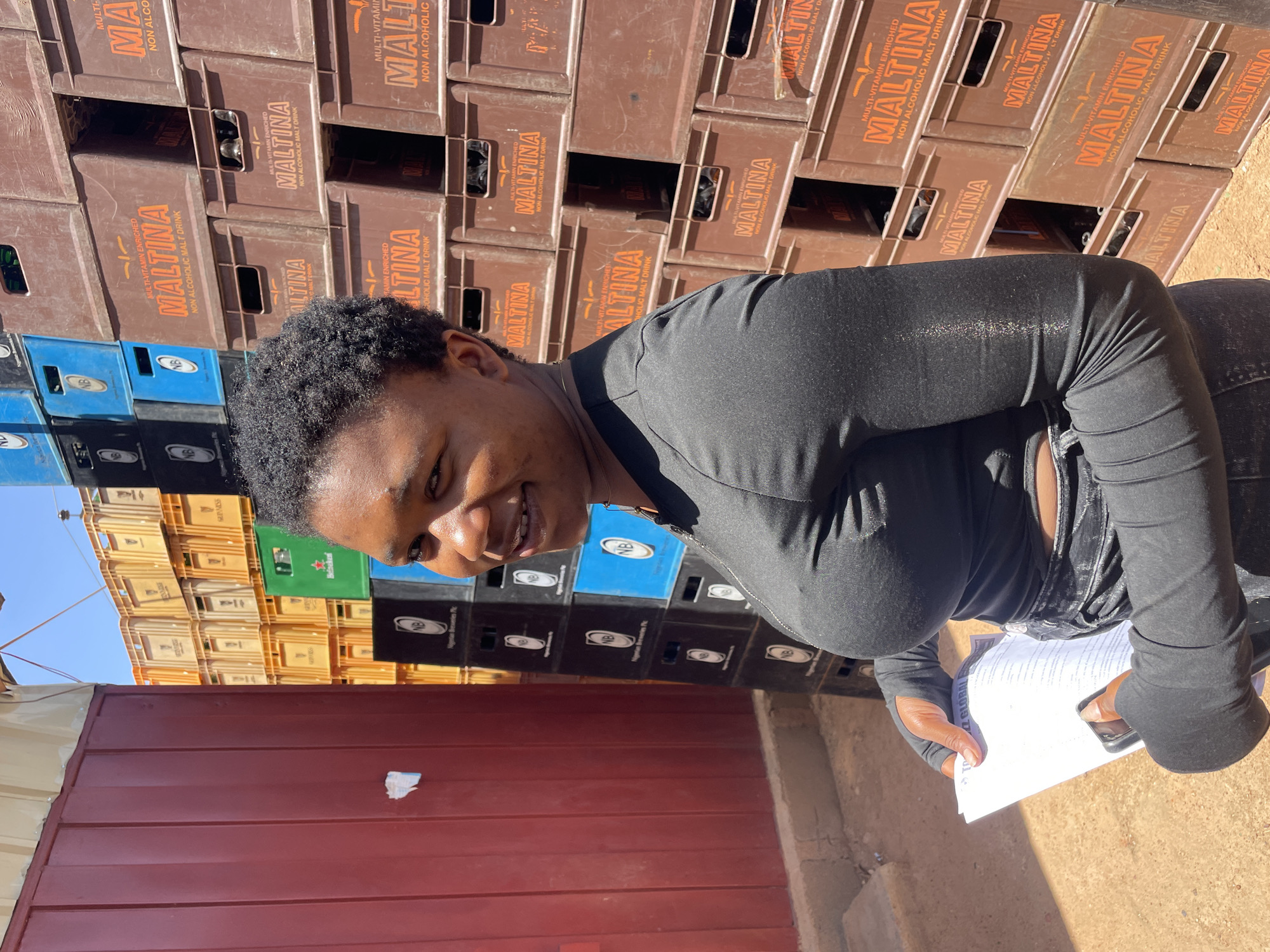 The photograph depicts a woman standing in what appears to be an outdoor industrial or storage area in Gura Top, Nigeria. The background consists of a stacked wall of brown plastic crates labeled "MALTINA NON ALCOHOLIC MALT DRINK," intermixed with crates of various brands and colors. The woman is positioned in front of the crate wall, holding a document in one hand and a mobile phone in the other. She is wearing a black, long-sleeved top and dark pants. A segment of a red corrugated metal wall is visible along the bottom edge of the image. The lighting indicates a sunny day.