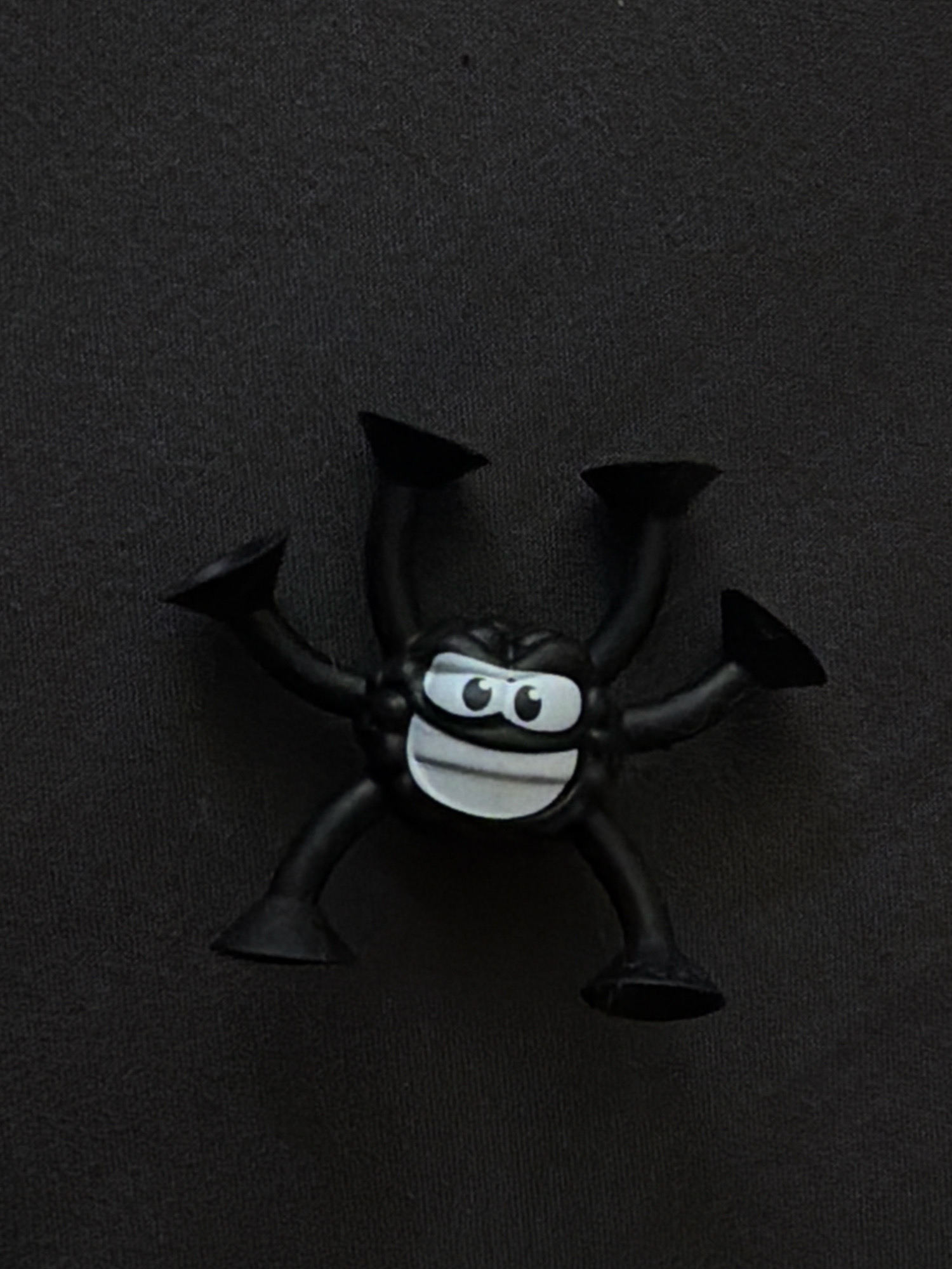 A black, multi-limbed toy with suction cups on the ends of its limbs is positioned centrally on a dark, textured surface. The toy has a face with large, white eyes with black pupils and a wide, smiling mouth with white teeth and a black outline. The toy is positioned as if it is about to adhere to or has just detached from a surface. The background is uniformly dark and appears to be fabric. No specific location in Hrodna, Belarus, can be identified from the image.
