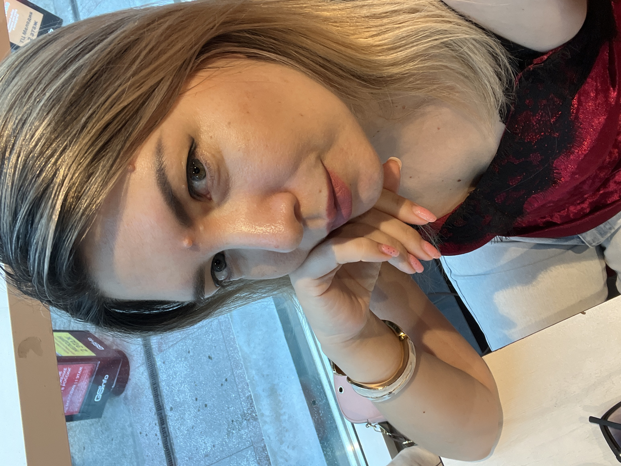 A woman with blonde hair and light skin, wearing a red top and white pants, poses for a selfie at a table in what appears to be an airport or train station in Sochi, Russia. She is resting her head on her hand, and her fingernails are painted pink. In the background, there is a window with a view of the exterior, and a sign with Cyrillic lettering can be seen through the glass. The lighting suggests it is daytime.