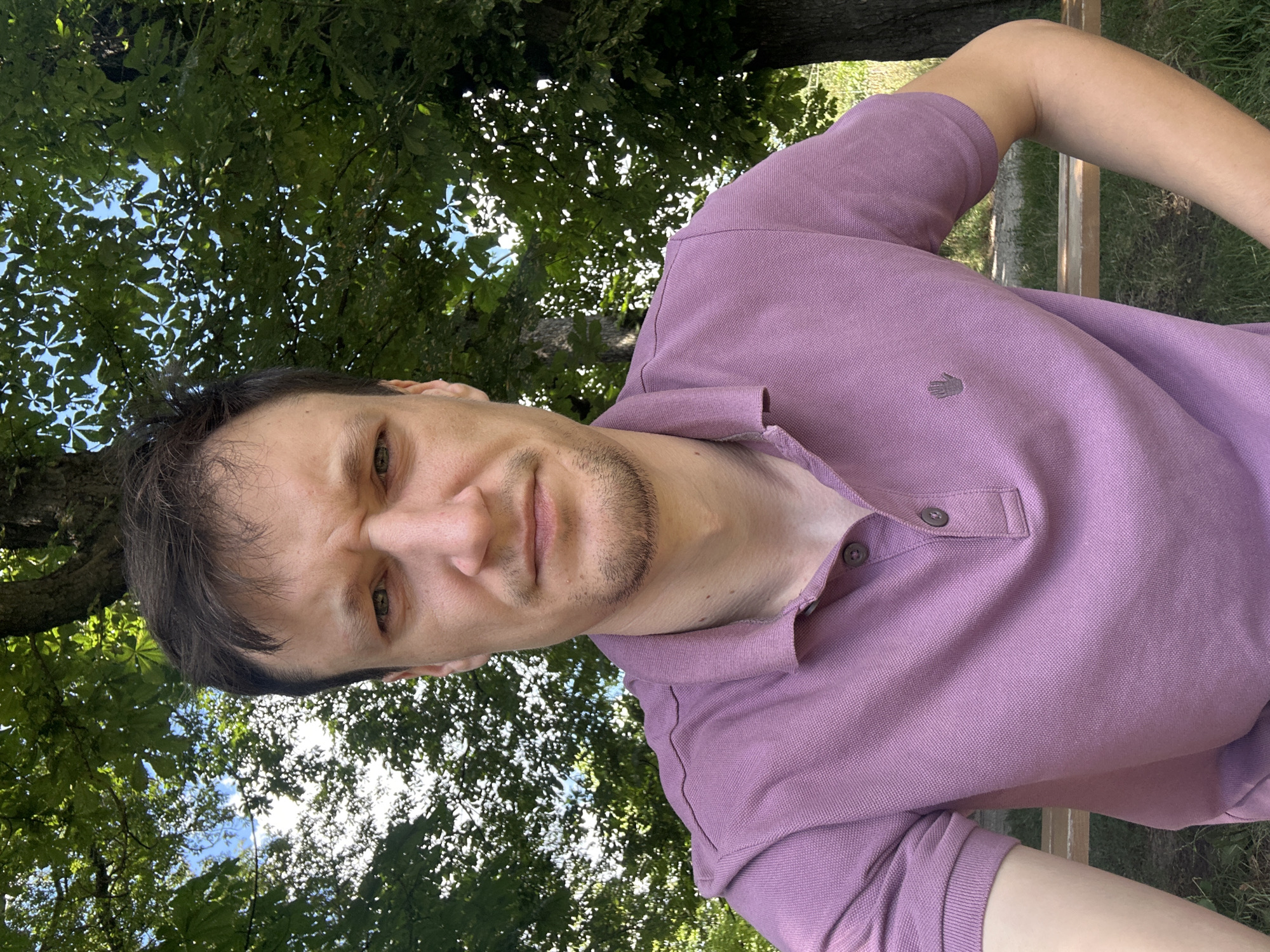 The image is a waist-up, selfie-style photograph of a light-skinned man with brown hair, wearing a short-sleeved, mauve polo shirt with two buttons at the neck. He has a stern expression with his eyebrows furrowed. The man is outdoors, seated on a wooden bench, with his left arm resting on his lap and his right hand on his hip. Behind him, out-of-focus, are green trees and dappled sunlight, suggesting a park or wooded area. A tree trunk is visible to the right of the man. The location is identified as Makiv, Ukraine.