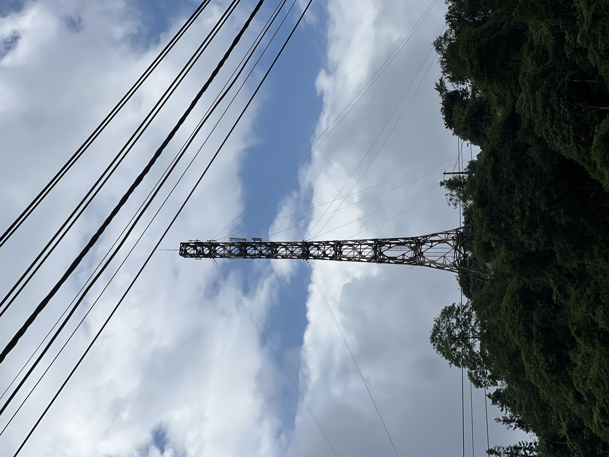 The image shows a tall, lattice telecommunications tower situated on a densely forested hillside. The tower is dark and metallic, with visible antennas and equipment. Several thick, dark power lines stretch diagonally across the upper portion of the frame, obscuring parts of the sky. The sky is partly cloudy, with patches of blue visible amongst the white and gray clouds. The hillside is covered in lush green trees and foliage, with the tower appearing to be built into the natural landscape. The scene is set in Quản Bạ Commune, Vietnam.