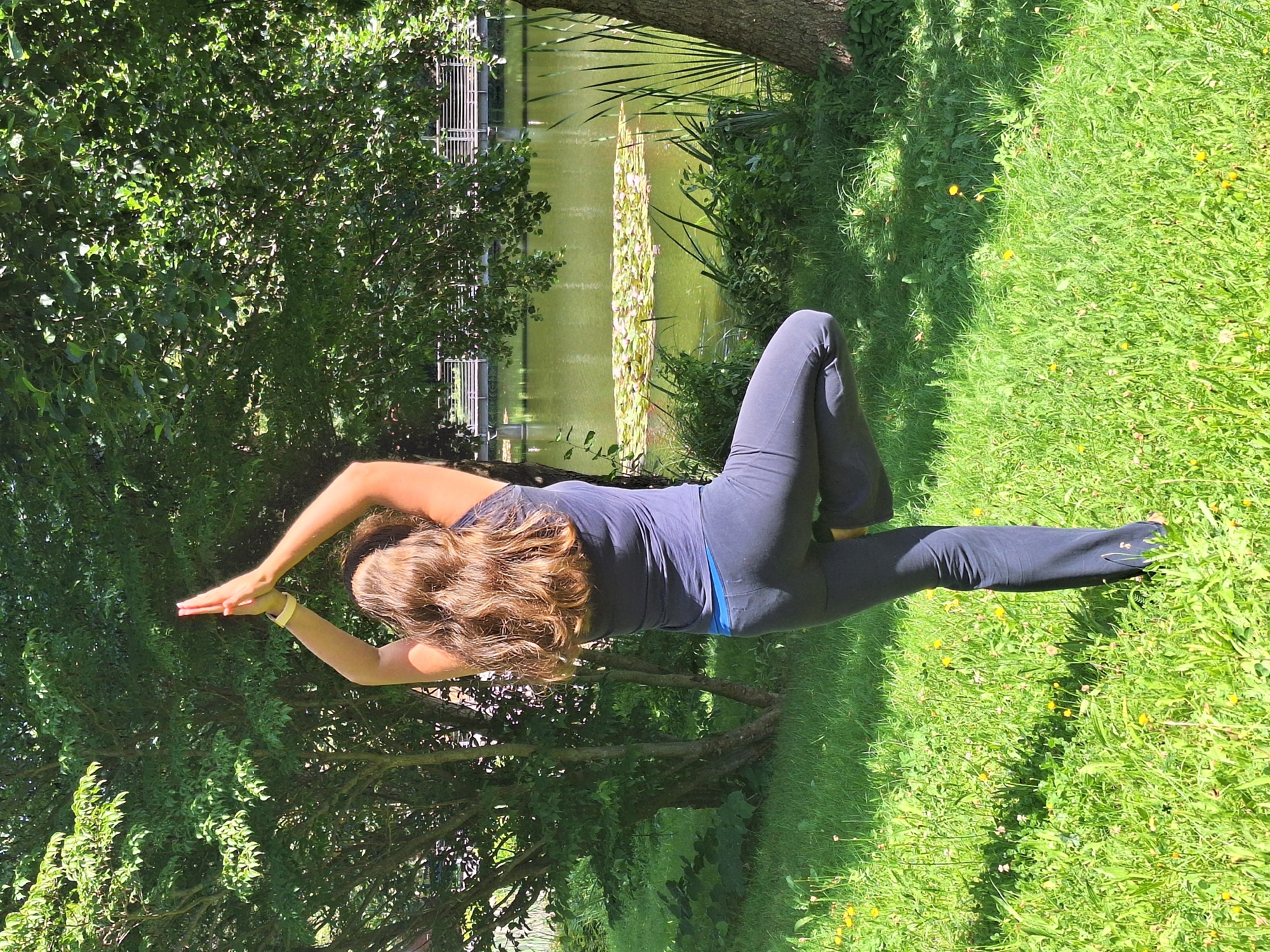 A person is practicing yoga in a grassy area next to a body of water. The person is seen from the back, facing away from the camera, with their arms raised above their head and hands pressed together. Their left leg is bent at the knee, with the foot resting on the inner thigh of the right leg, a pose commonly known as Tree Pose in yoga. The person is wearing dark athletic pants and a dark tank top. The environment features lush green grass dotted with small yellow flowers, surrounded by trees with dense foliage. In the background, a calm body of water with lily pads is visible, and a bridge or walkway can be seen across the water. The scene is illuminated by bright sunlight, casting shadows on the grass. This image appears to have been taken in a park or natural area. The city of Wolkenstein, Germany, is known for its natural beauty and recreational areas, making such a scene plausible within its surroundings.