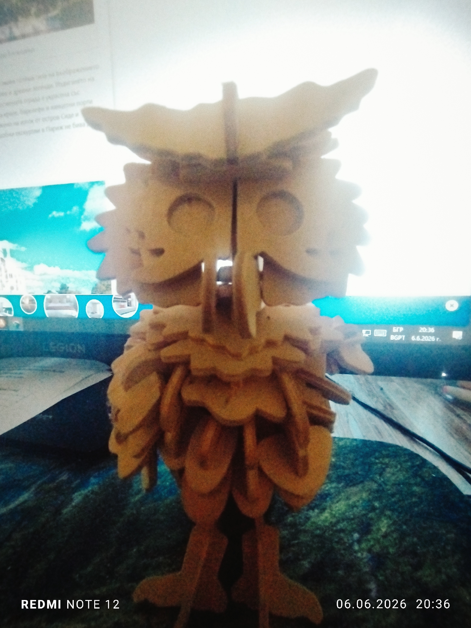 A forensic examination of the image reveals a detailed indoor scene centered around a three-dimensional wooden owl puzzle, captured from a slightly low-angle perspective. The environment appears to be a desk setting in Burgas, Bulgaria, based on contextual clues.
**Subject:**
The primary subject is a meticulously constructed wooden puzzle model of an owl, standing upright in the foreground. The owl is assembled from multiple interlocking, light-brown wooden pieces, creating a layered, textured effect reminiscent of feathers. It features distinct pointed ears, two prominent circular eye sockets, a small beak-like protrusion, and sturdy, flat rectangular legs with broad feet.
**Environment and Objects:**
The wooden owl rests on a flat surface, likely a desk, which is covered by a patterned mat or mousepad displaying a dark, abstract green and blue design that resembles moss or a textured landscape. A thin, black cable extends across the right side of this surface. In the background, a computer monitor is prominently displayed.
**Computer Screen Details:**
The monitor's screen shows an active display. The upper-left portion contains text in Cyrillic script, with specific words like "Париж" (Paris) being legible, suggesting the content is a document or webpage in Russian or Ukrainian. Below this text, a bright blue sky with white clouds is visible, possibly part of a background image or video. A horizontal banner with several circular icons is present further down. The lower-left bezel or a panel beneath the screen displays a partial "LEGION" logo.
The lower-right corner of the monitor screen displays system information: "БГР" (BGR), identifying the location as Bulgaria; the current time "20:36"; and the date "6.6.2026 г." (year), accompanied by the text "BGPT".
**Metadata:**
The photograph was captured by a "REDMI NOTE 12" device, as indicated by the watermark in the bottom-left corner of the image. A timestamp "06.06.2026 20:36" is present in the bottom-right corner, corroborating the date and time displayed on the computer monitor. The "БГР" (BGR) indicator on the screen firmly places the location within Bulgaria, aligning with the requested detail of Burgas.