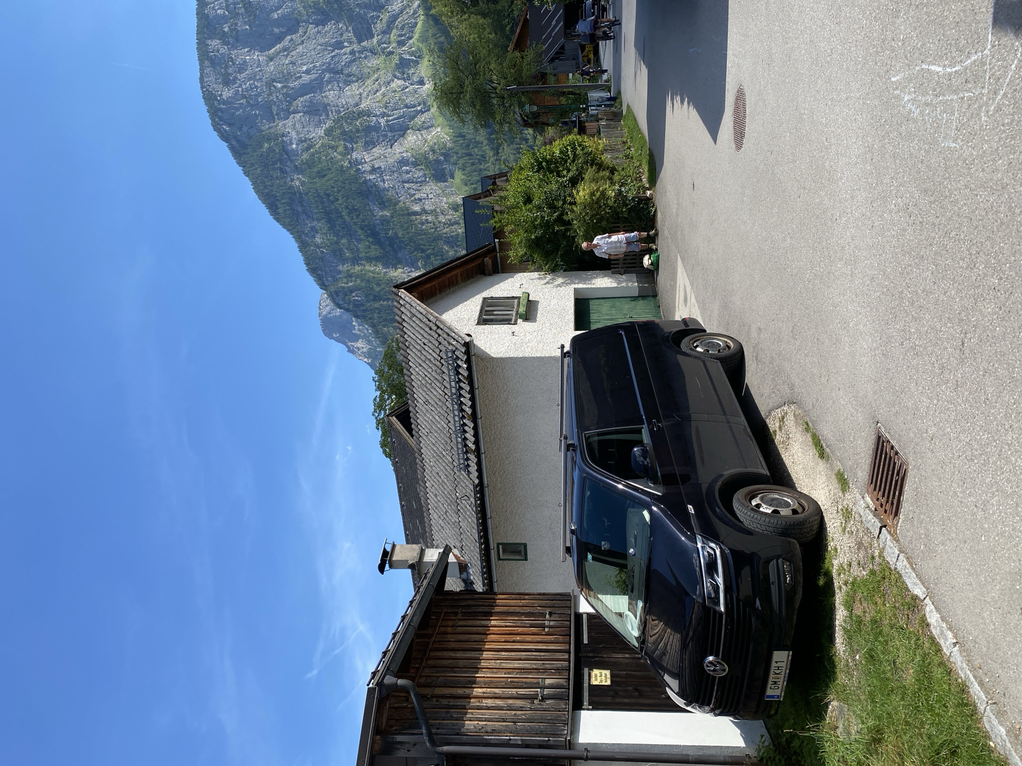 A black Volkswagen Transporter van is parked on the left side of a narrow, paved street in Hallstatt, Austria. The van has a roof rack and an Austrian license plate that reads "GM KH 1". To the left of the van is a wooden-clad building with a white foundation and a gray-tiled roof. Next to it is a white stucco house with a gray-tiled roof and a green-trimmed window. A man in a plaid shirt and shorts stands next to the white house. Further down the street, on the right, several people are cycling and a car is parked. Behind the buildings, a steep, rocky mountain covered in trees rises dramatically under a clear blue sky. The scene is illuminated by bright sunlight, casting shadows on the street and buildings.