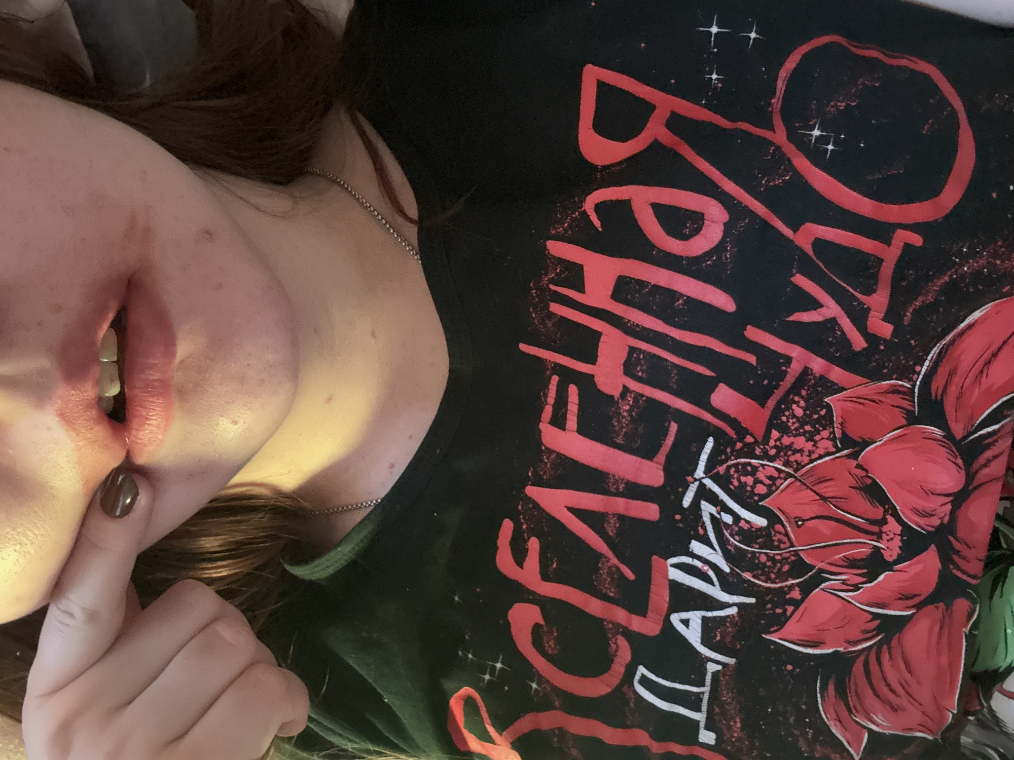 A person is visible from the chest up, lying on their back. They are wearing a black t-shirt with red and white lettering and a rose graphic. The person's left hand is raised near their face, with a finger touching their lips. They are wearing a silver chain necklace. The person has brown hair. The image does not show any evidence of being taken in Izhevsk, Russia, or any location information.