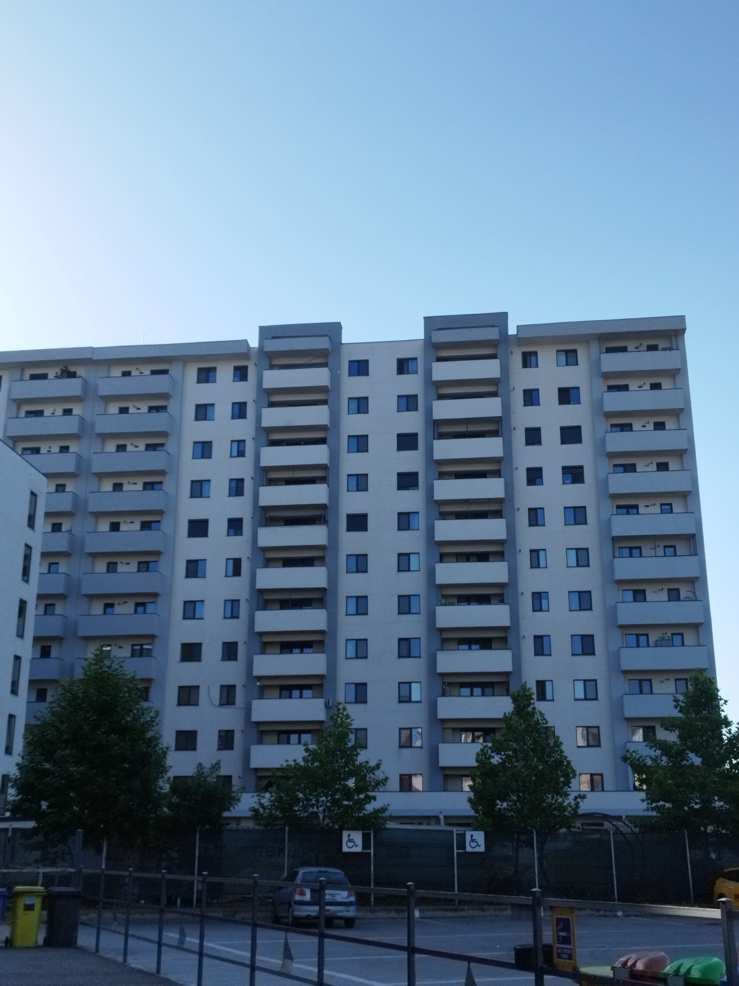 The image shows a large apartment building with multiple balconies, set against a clear blue sky. The building is light gray with darker gray accents around the balconies and on the roofline. Numerous windows are visible on each floor. In front of the building, there is a row of trees and a fence. Below the fence, a street or parking area is visible with a car, two trash cans, and a sign indicating a disabled parking spot. The scene appears to be in an urban environment, likely a residential area. The specific location is Bucharest, Romania, as indicated by the text on a yellow sign which reads "PARCARE PERSOANE CU HANDICAP" (Parking for people with disabilities).